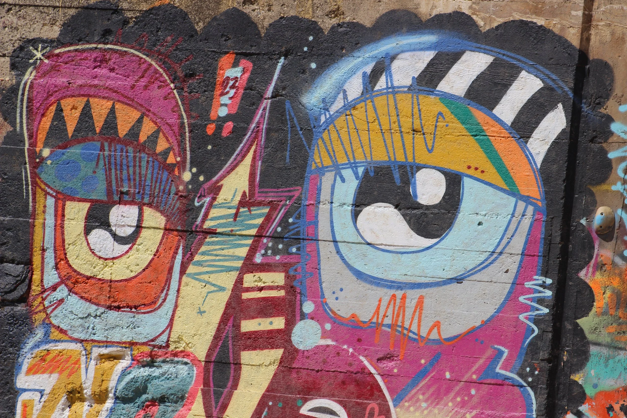 Graffiti mit zwei großen, stilisierten Anime-Augen, bunten geometrischen Formen und Mustern auf einer Wand.