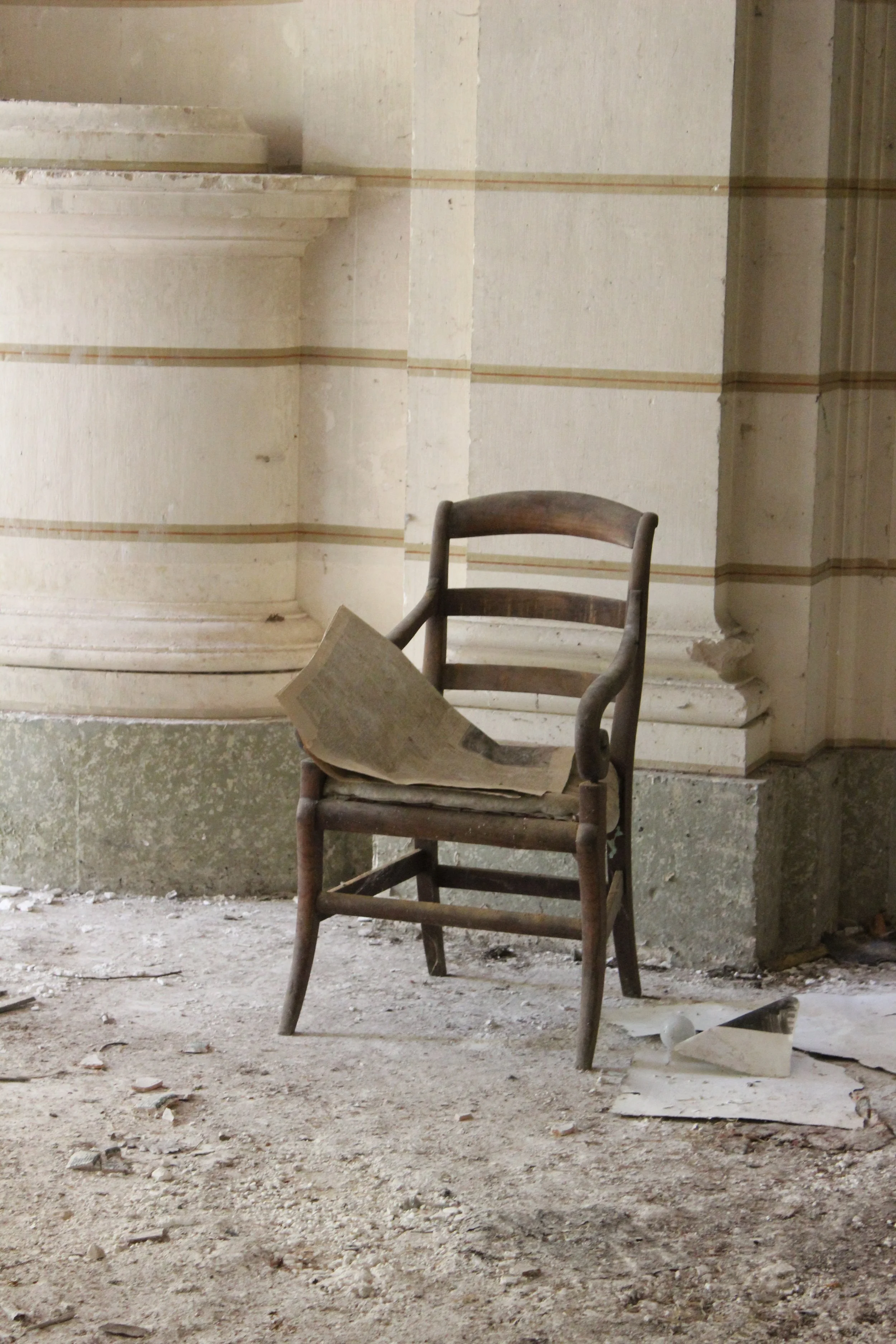 Ein alter Holzzaunstuhl mit einem aufgeschlagenen Buch liegt in einem staubigen, verlassenen Raum mit beschädigten Wänden und zerbrochenem Glas auf dem Boden.