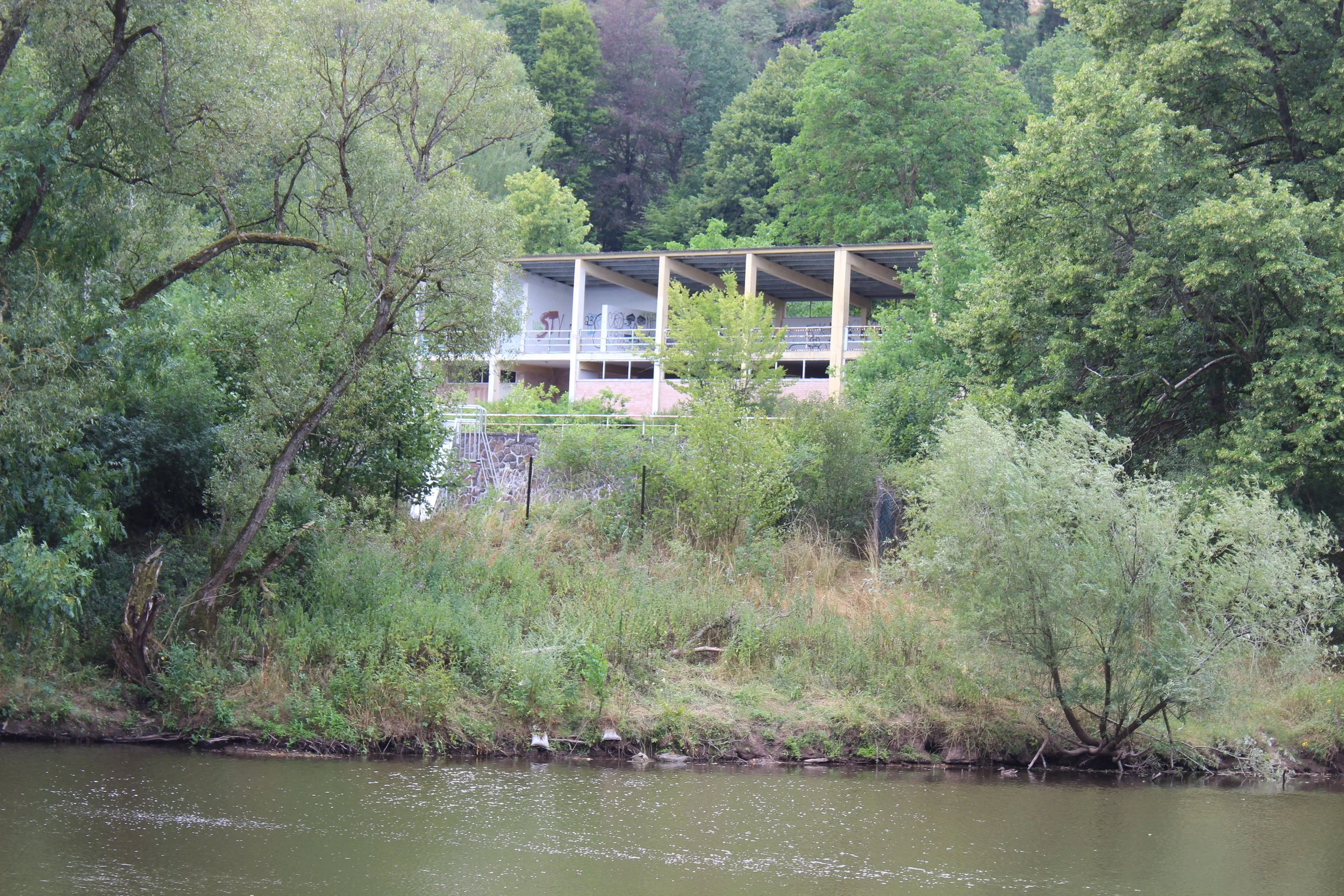 Ein verlassenes, verfallenes Gebäude auf einem Hügel, umgeben von Bäumen und dichtem Grün, in der Nähe eines Flusses.