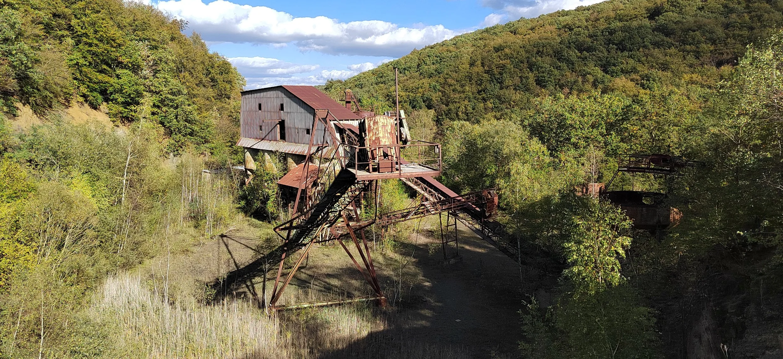 Verrostete, alte Bergwerksanlage in einer bewaldeten Hügellandschaft bei Sonnenlicht