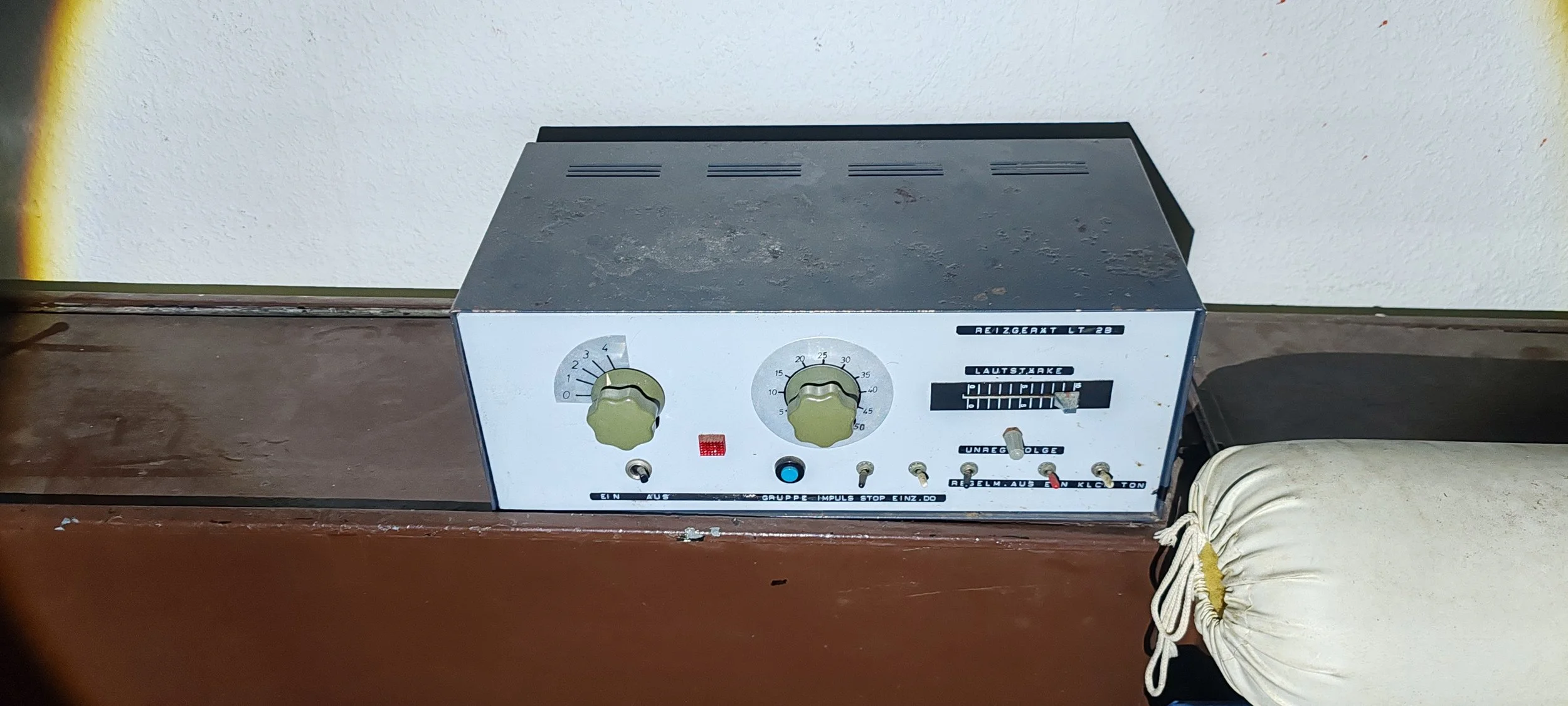 Ein altmodisches, statisches Elektrogerät mit zwei Einstellknöpfen, mehreren Schaltern und einem Lautstärkenregler, auf einer Holztischplatte, neben einem weißen gefalteten Kissen.