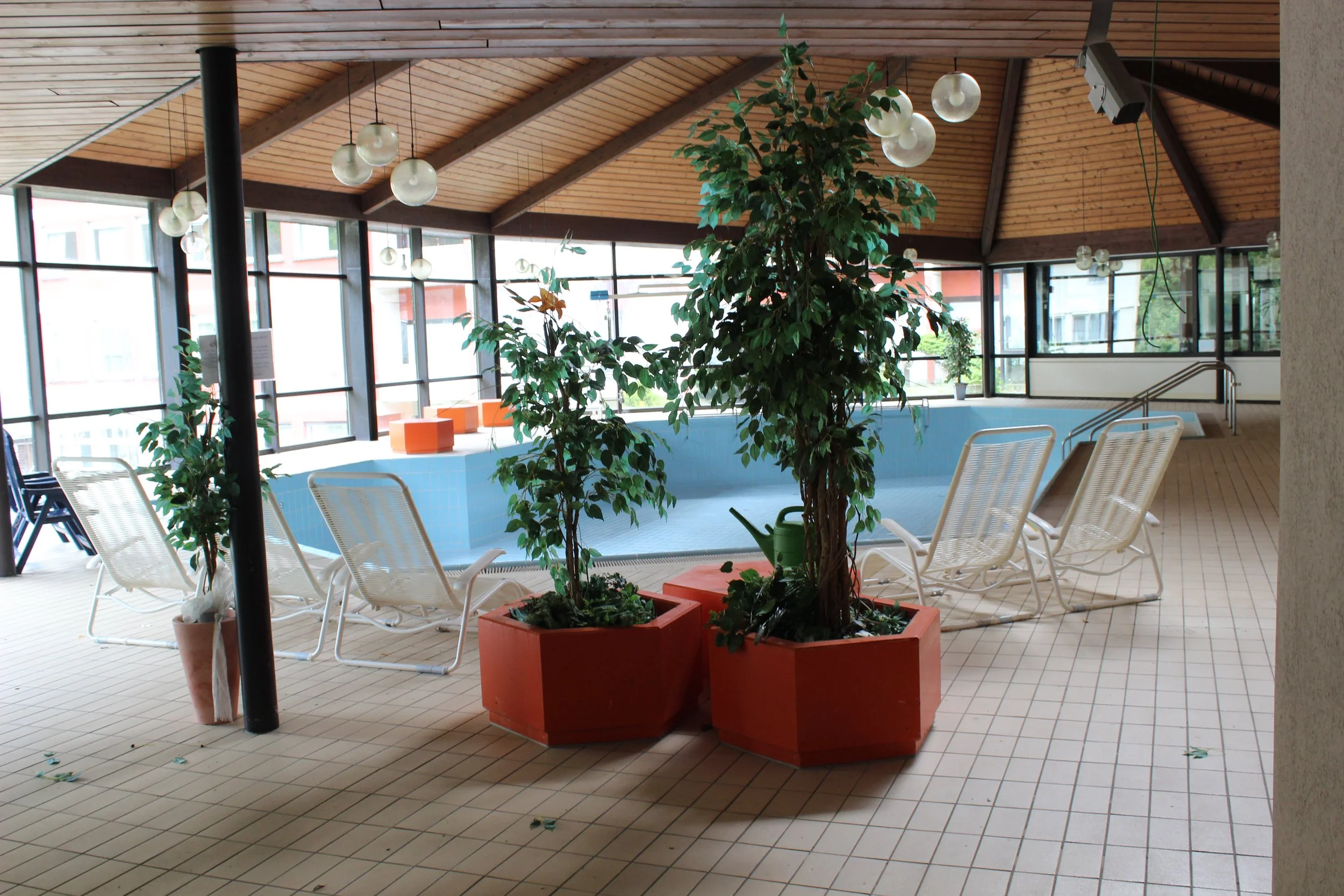 Innenraum eines Wellnessbads mit Pool, Liegestühlen, Pflanzen in großen Töpfen und hölzernem Dach.