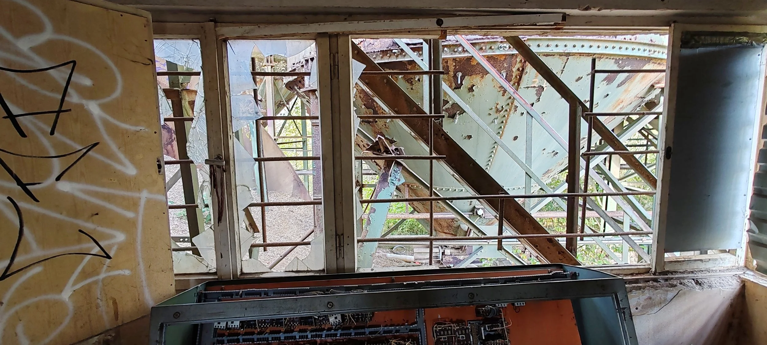 Blick durch ein zerbrochenes Fenster auf eine rostige Metallstruktur im Freien, mit sichtbaren Schrauben und Rahmen