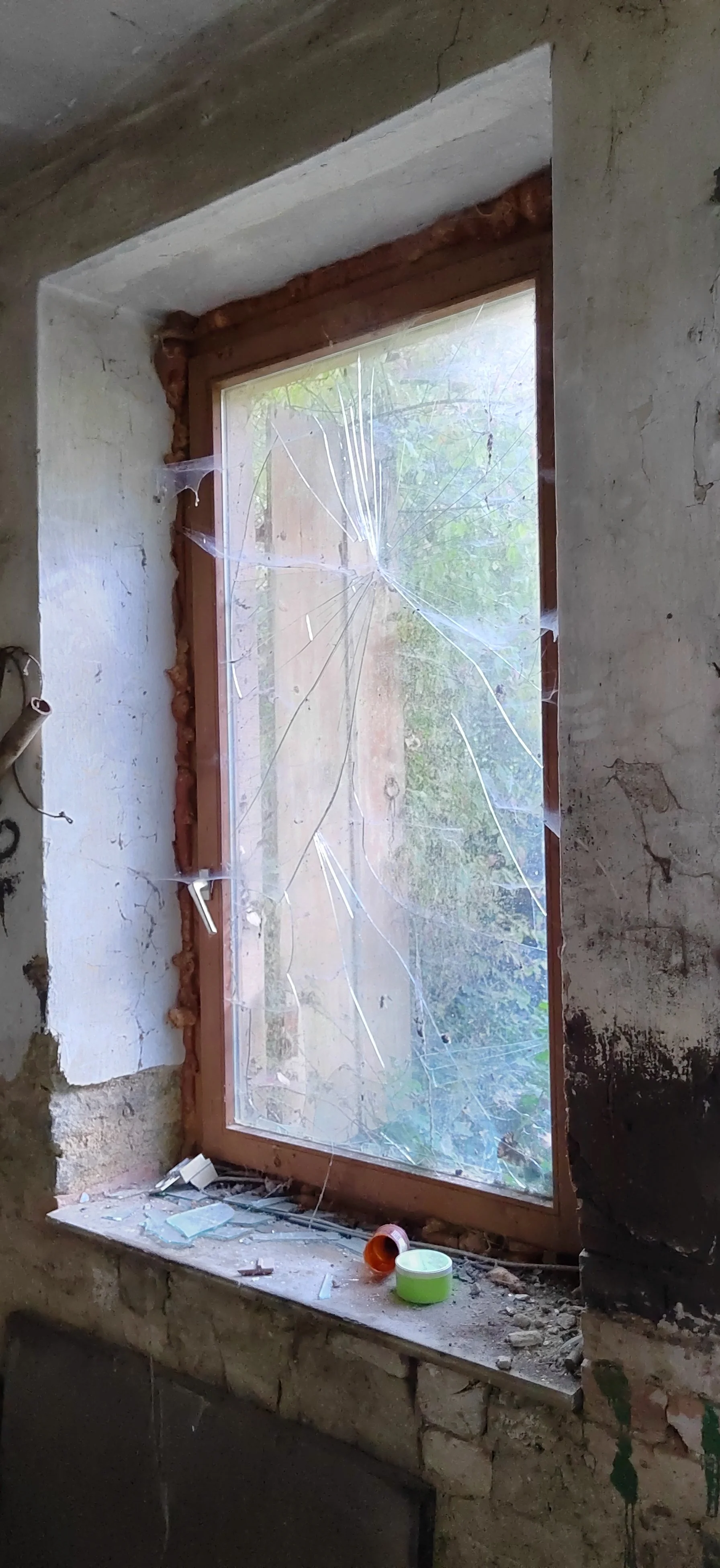 Ein zerbrochenes Fenster mit Spinnweben in einem alten Haus, umgeben von Bau-Resten.
