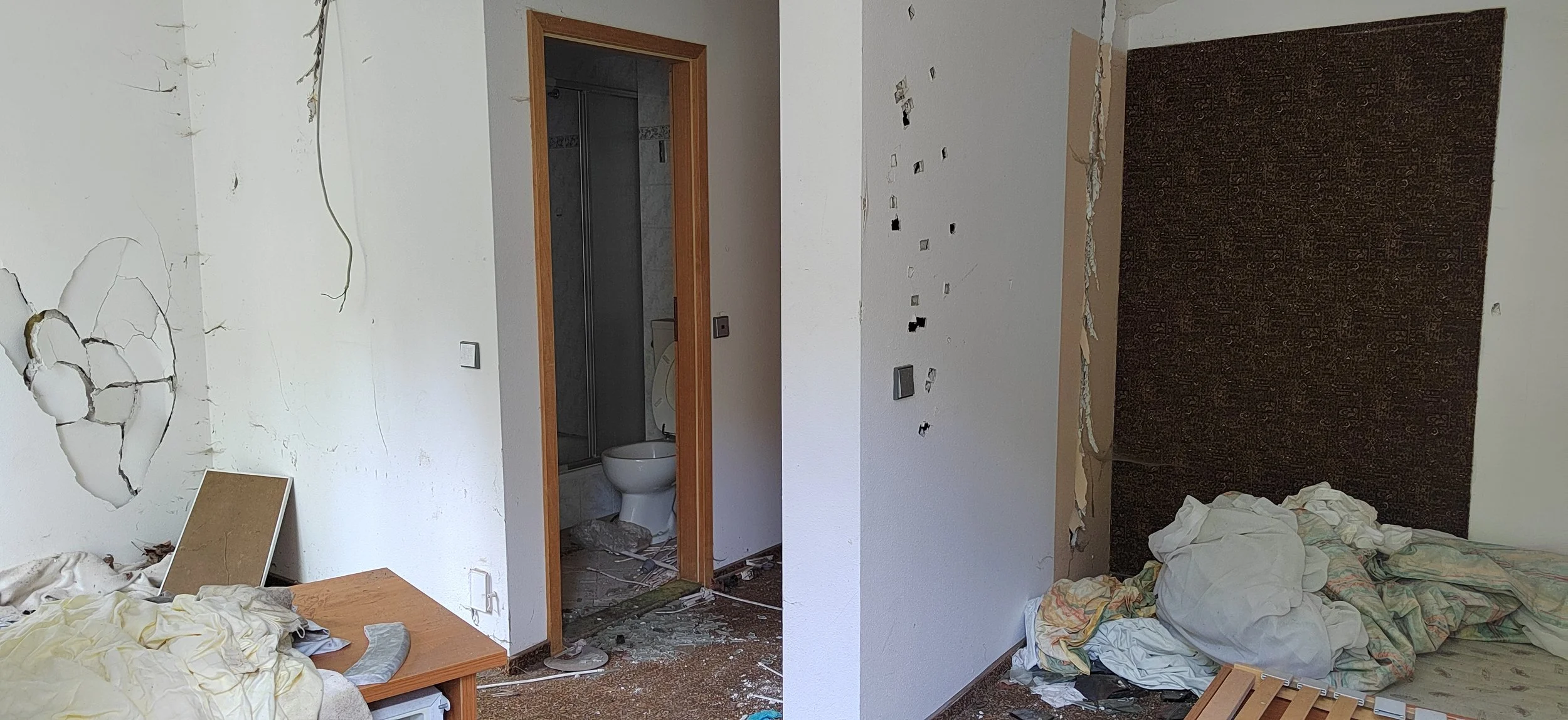 Zimmer mit beschädigten Wänden, einem unordentlichen Bett, einem Nachttisch, einem offenen Badezimmer mit WC, zerbrochenen Gegenständen auf dem Boden und einem chaotischen Raum.