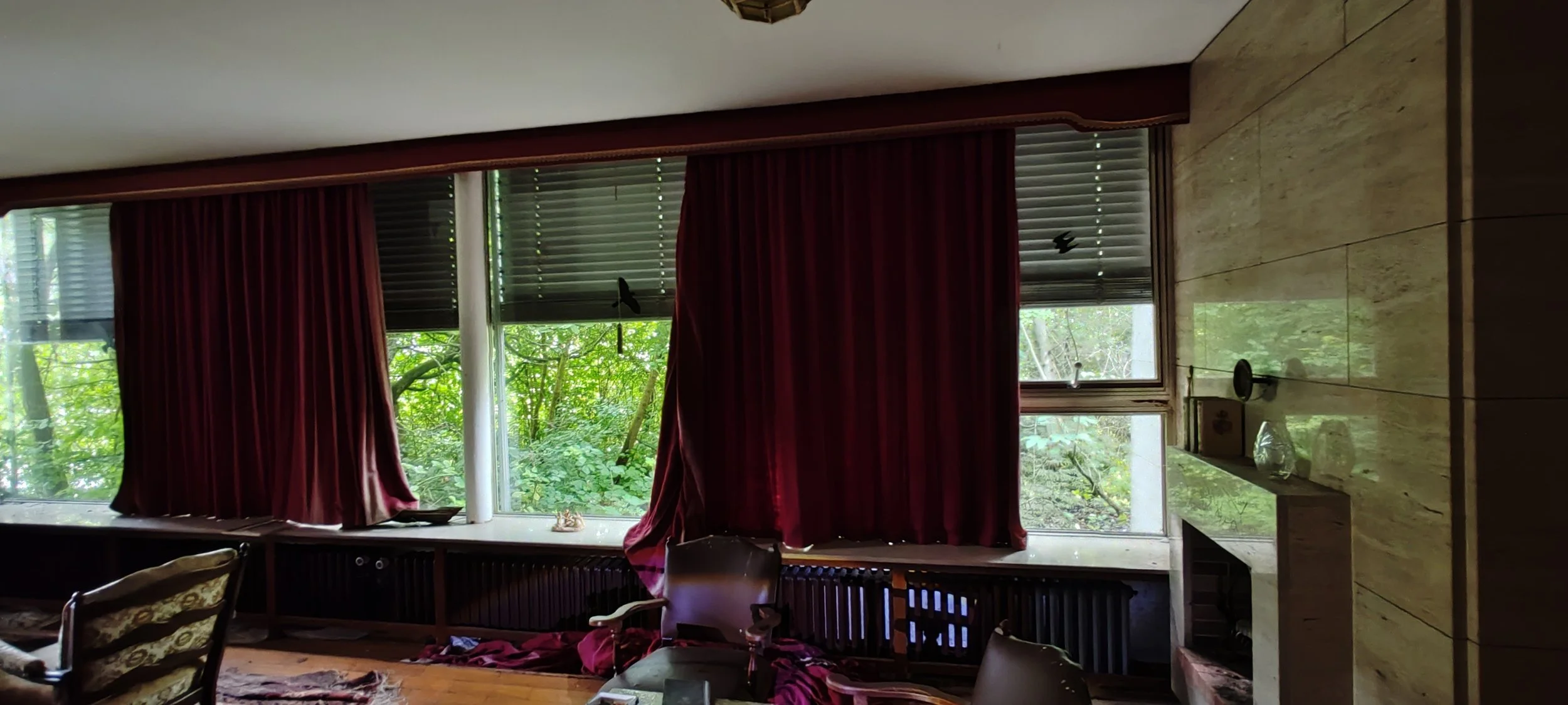 Ein Raum mit großen Fenstern, dunklen roten Vorhängen, und grüner Natur draußen sichtbar, einige Möbel und Dekor auf einer Fensterbank.