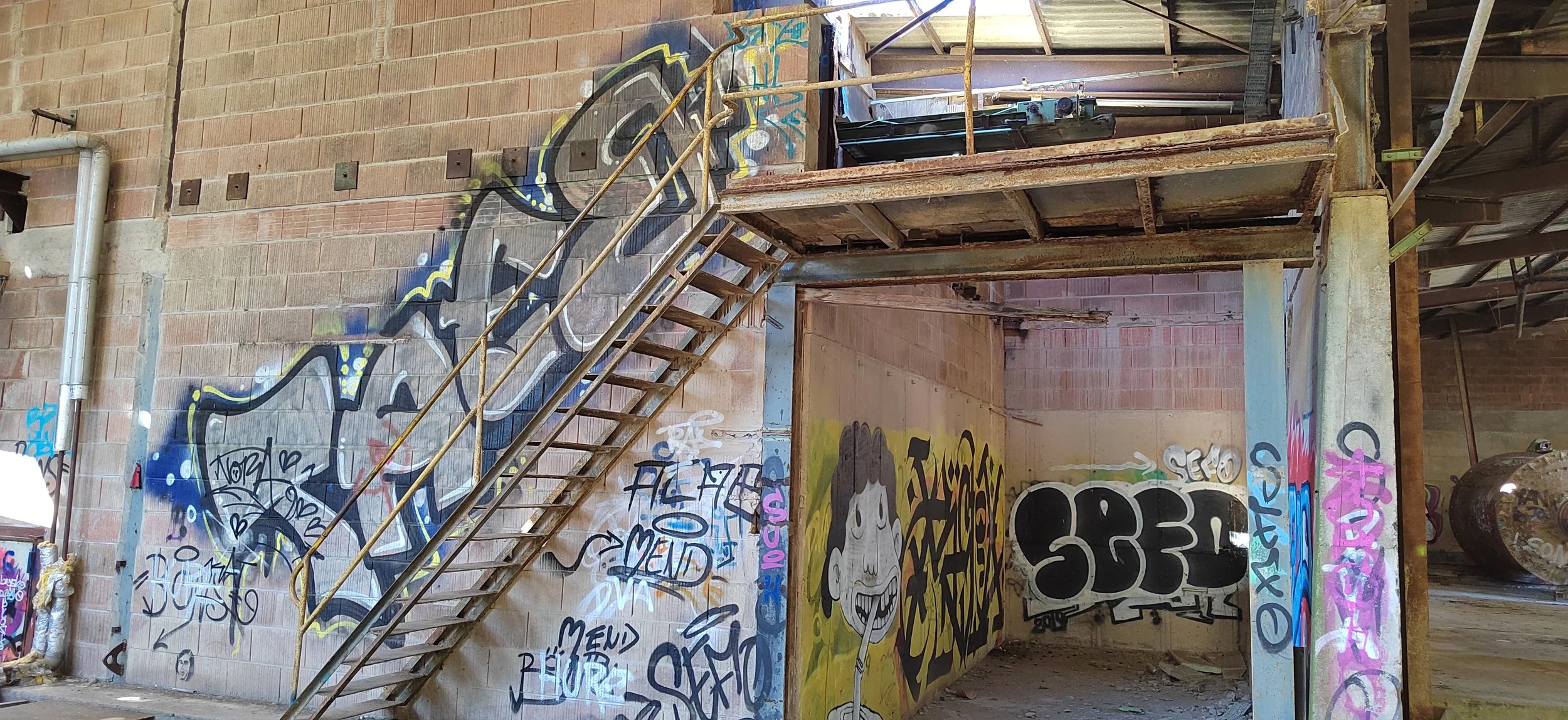 Innerhalb eines verfallenen Gebäudes mit grauer Betonwand, die mit Graffiti bedeckt ist, eine schmale Treppe aus Holz und Metall führt zu einer höheren Ebene. Es gibt ein großes Graffiti-Bild eines lachenden Gesichts, das auf einer Wand gemalt ist, s