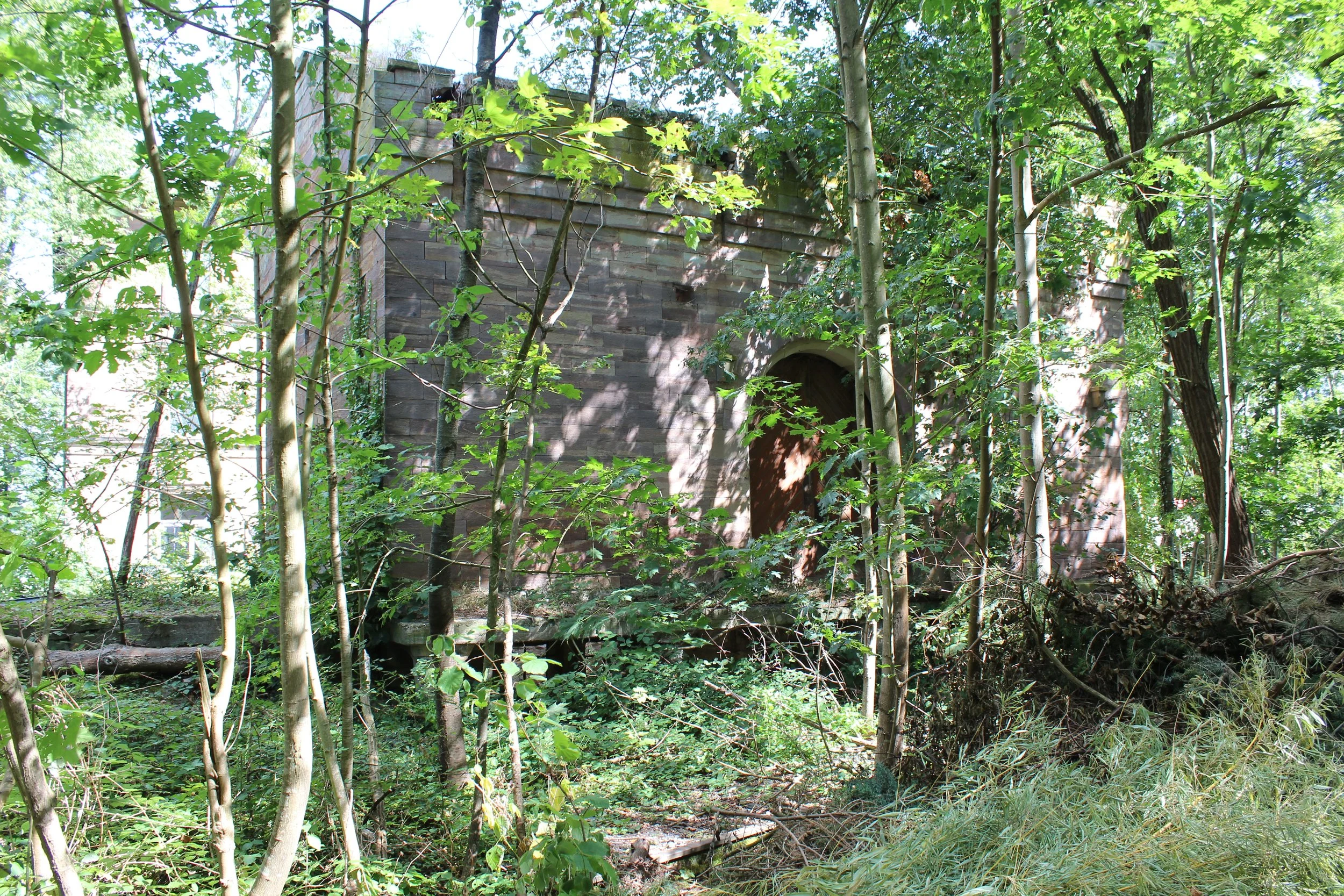Verfallenes Gebäude im Wald, umgeben von Büschen und Bäumen, mit einer vermodernden Fassade und einem Bogen am Eingang