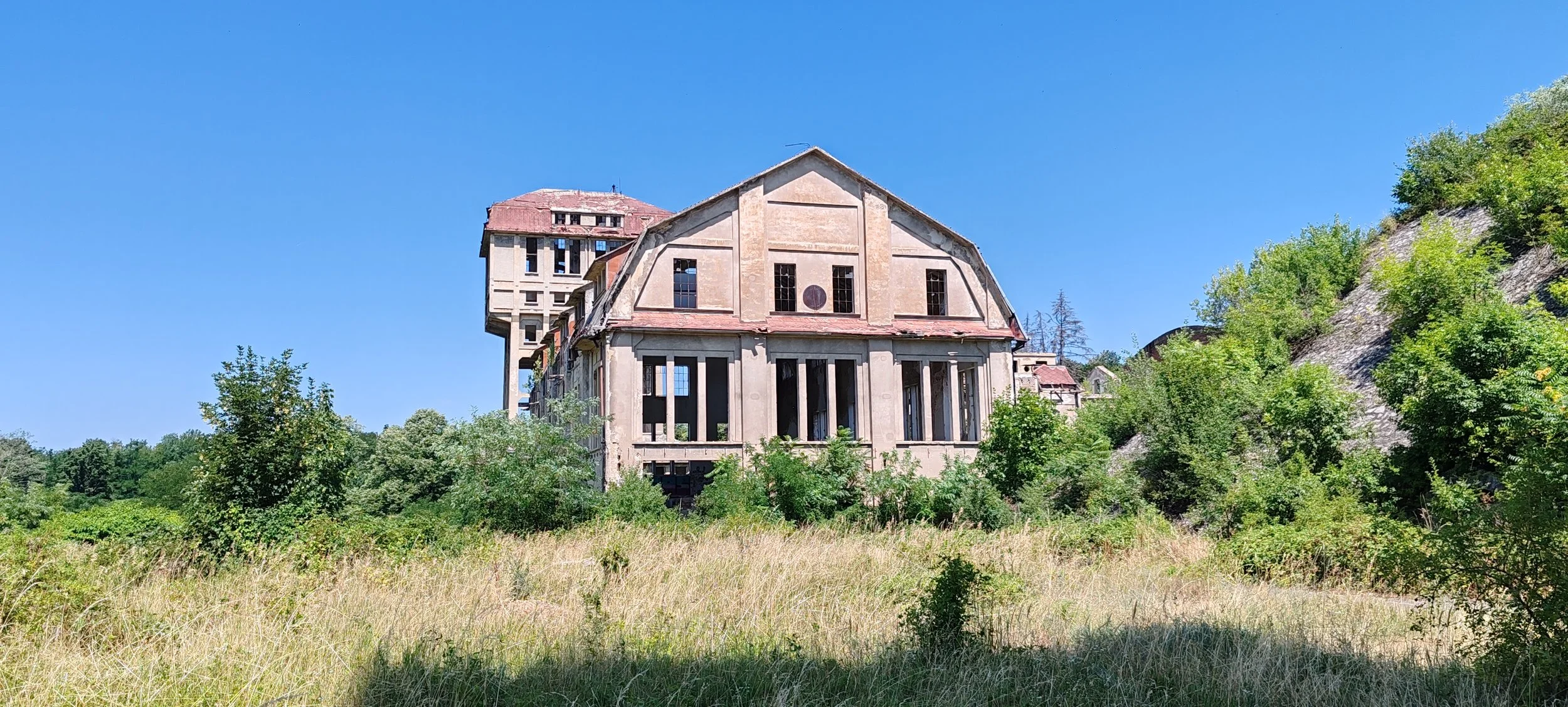 Verfallenes, großes Haus auf einem Hügel, umgeben von Bäumen und hohem Gras, unter klarem blauen Himmel.