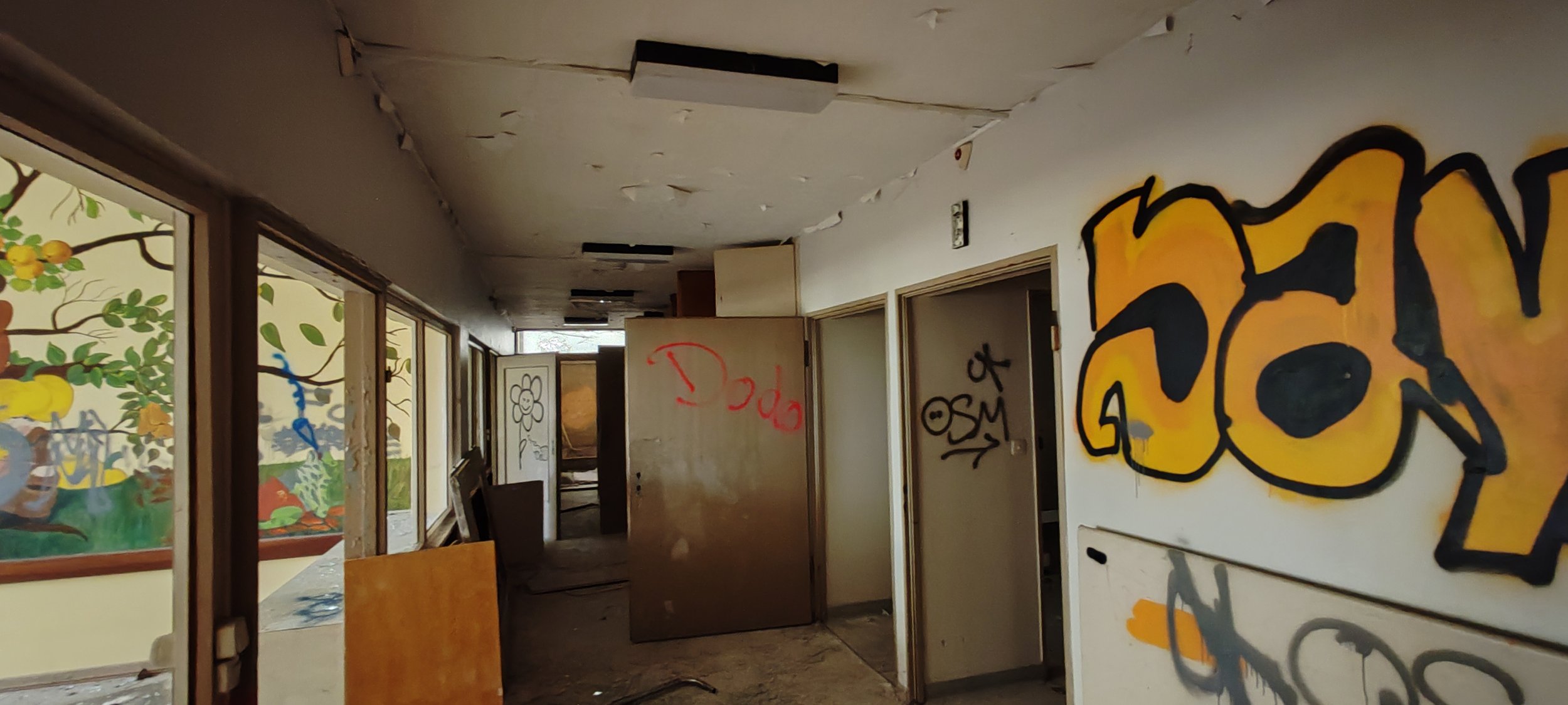 Verlassenes Gebäude mit grauen, abgenutzten Wänden, Graffiti und zerbrochenen Fenster. Innen ist es chaotisch mit umherliegenden Gegenständen und kaputtem Mobiliar.