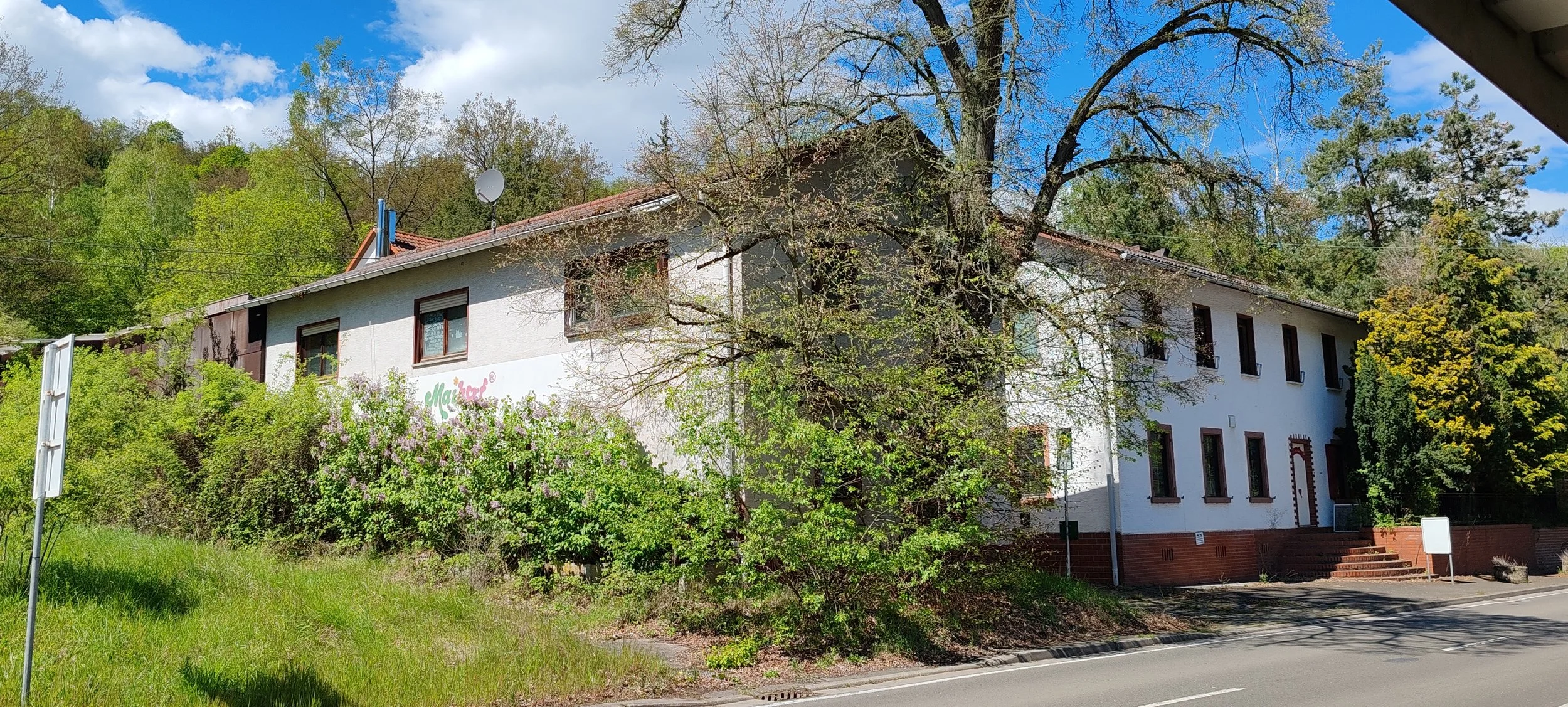 Ein weißes, mehrstöckiges Gebäude mit roten Ziegel-Details an einem sonnigen Tag, umgeben von Bäumen und Sträuchern, mit einer Straße im Vordergrund.