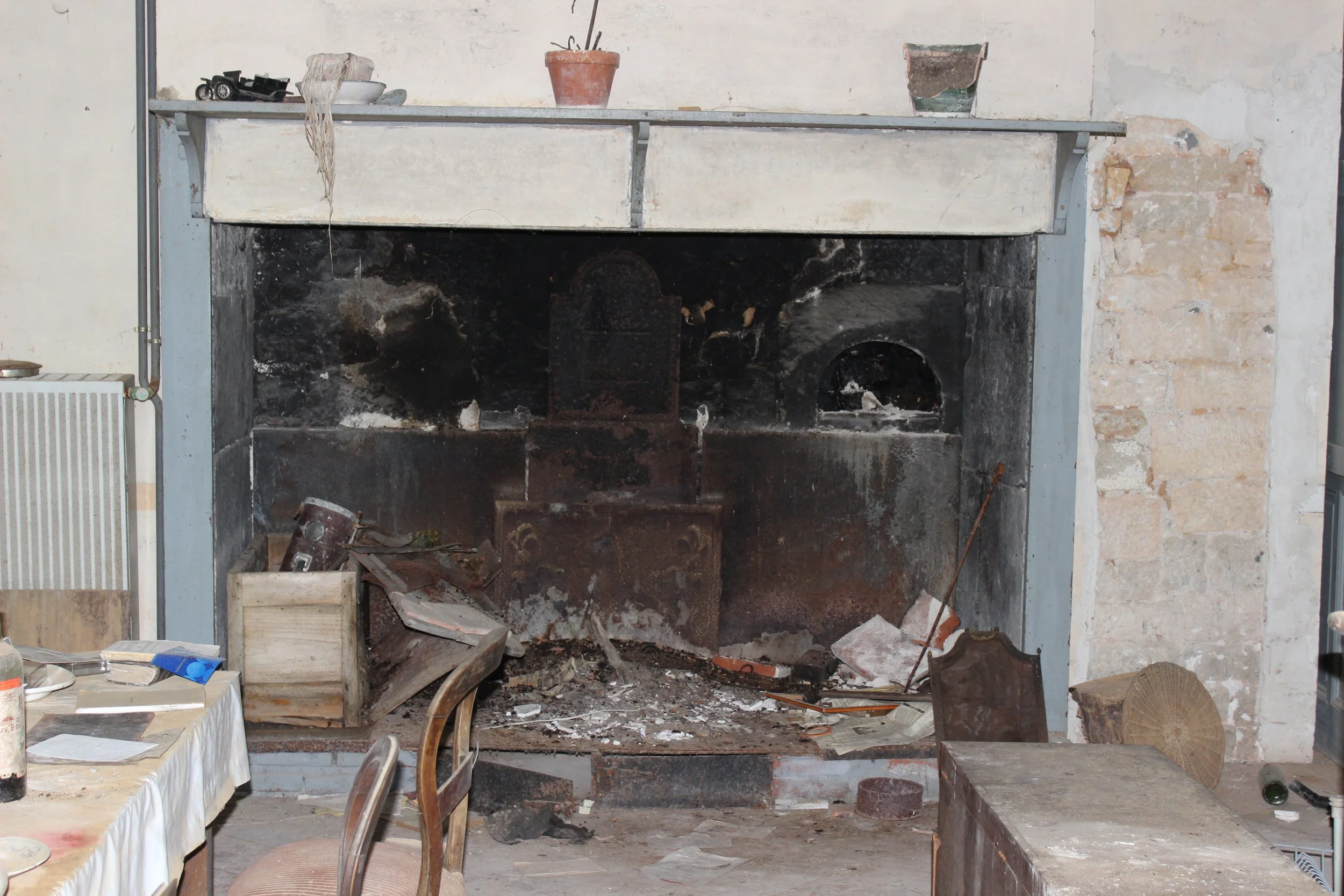 Ein alter, verfallener Kamin mit Ruß und Ascheresten. Im Raum steht auch ein Tisch mit Stühlen und verschiedenen Gegenständen, alles wirkt ungepflegt und vernachlässigt.
