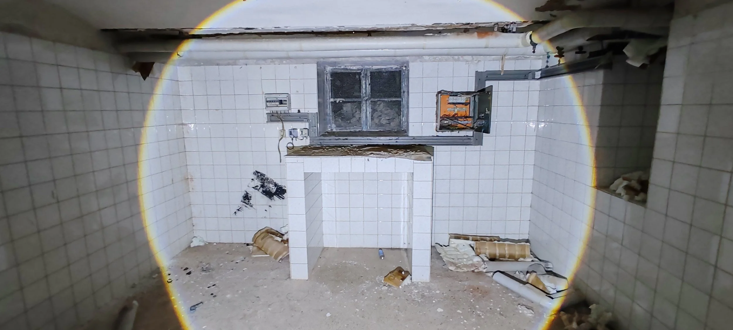 Ein verlassener Raum mit weißen Fliesen, einer kleinen Theke, beschädigten Wänden und einem Fenster mit Gitter. Der Boden ist schmutzig und mit zerbrochenem Material bedeckt.