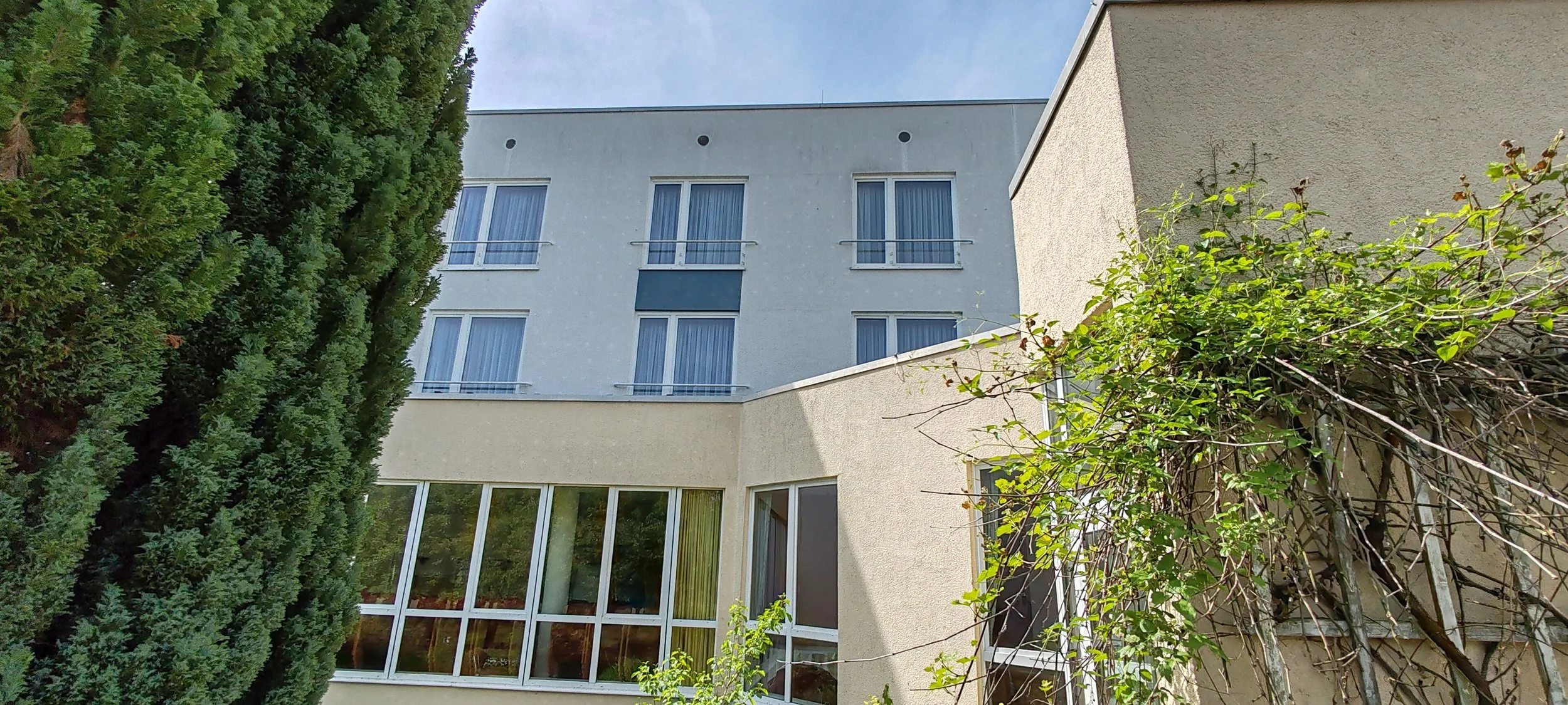 Mehrstöckiges Gebäude mit großen Fenstern, umgeben von Bäumen und Sträuchern, bei sonnigem Himmel.