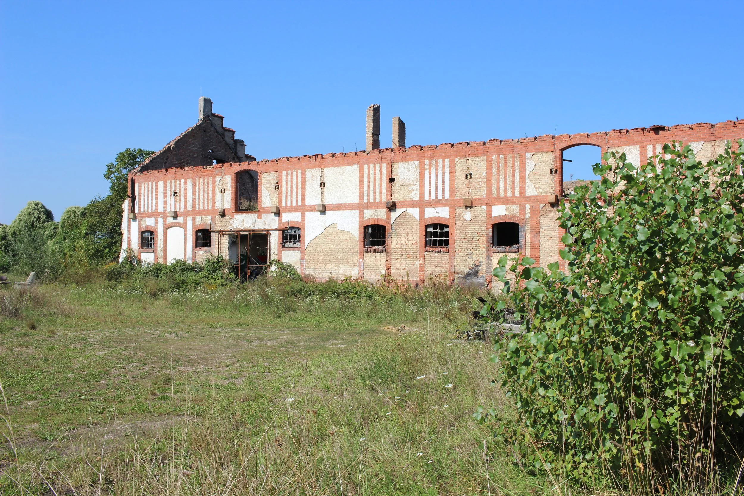 Verfallenes, halb zerfallenes Backsteingebäude mit Ziegelmauerwerk und Fenstern, umgeben von unkrautbedecktem Gelände, bei sonnigem Wetter.