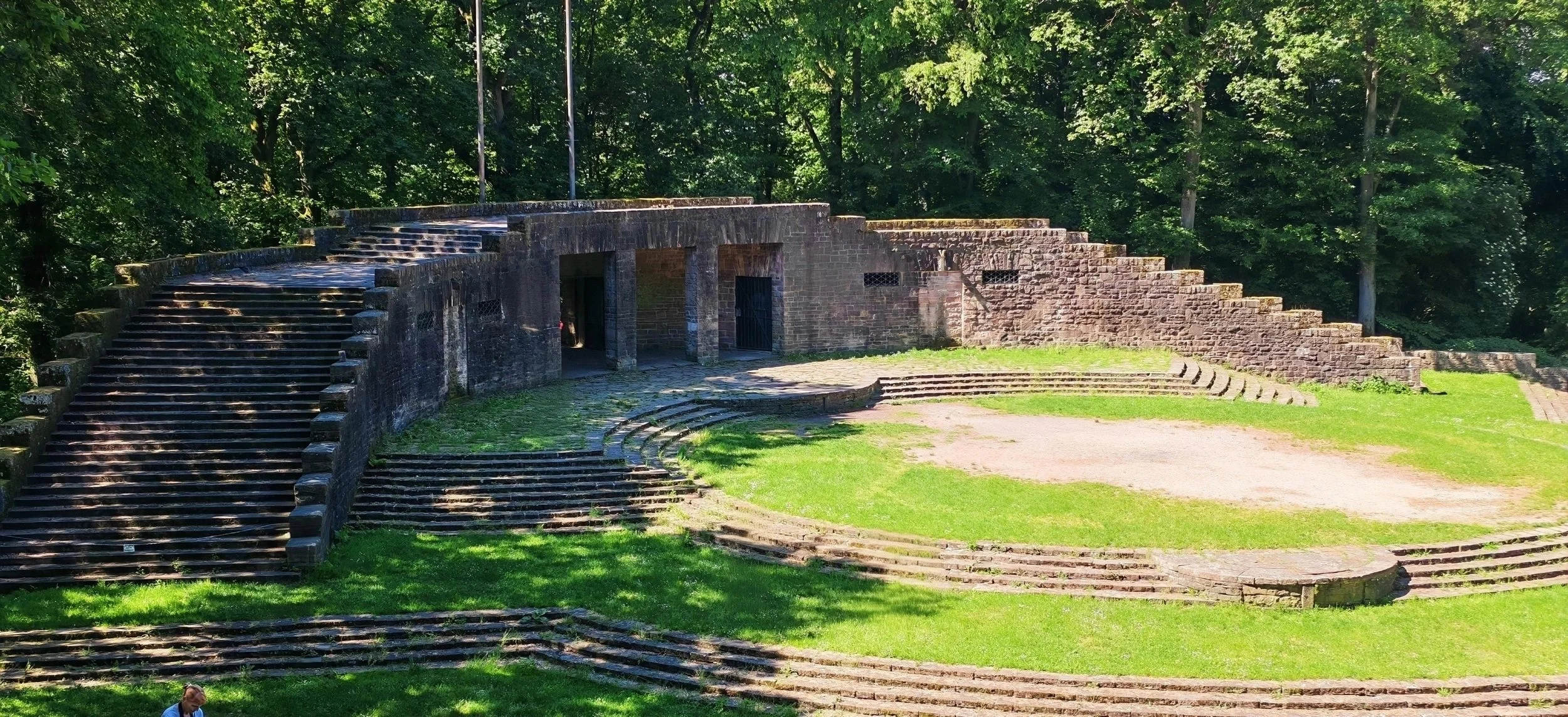 Verfallene Freilichtbühne aus Stein, eingebettet in einen grünen Park mit Bäumen im Hintergrund. Die Bühne hat alte Treppen und eine offene Nähe mit zwei Türen.