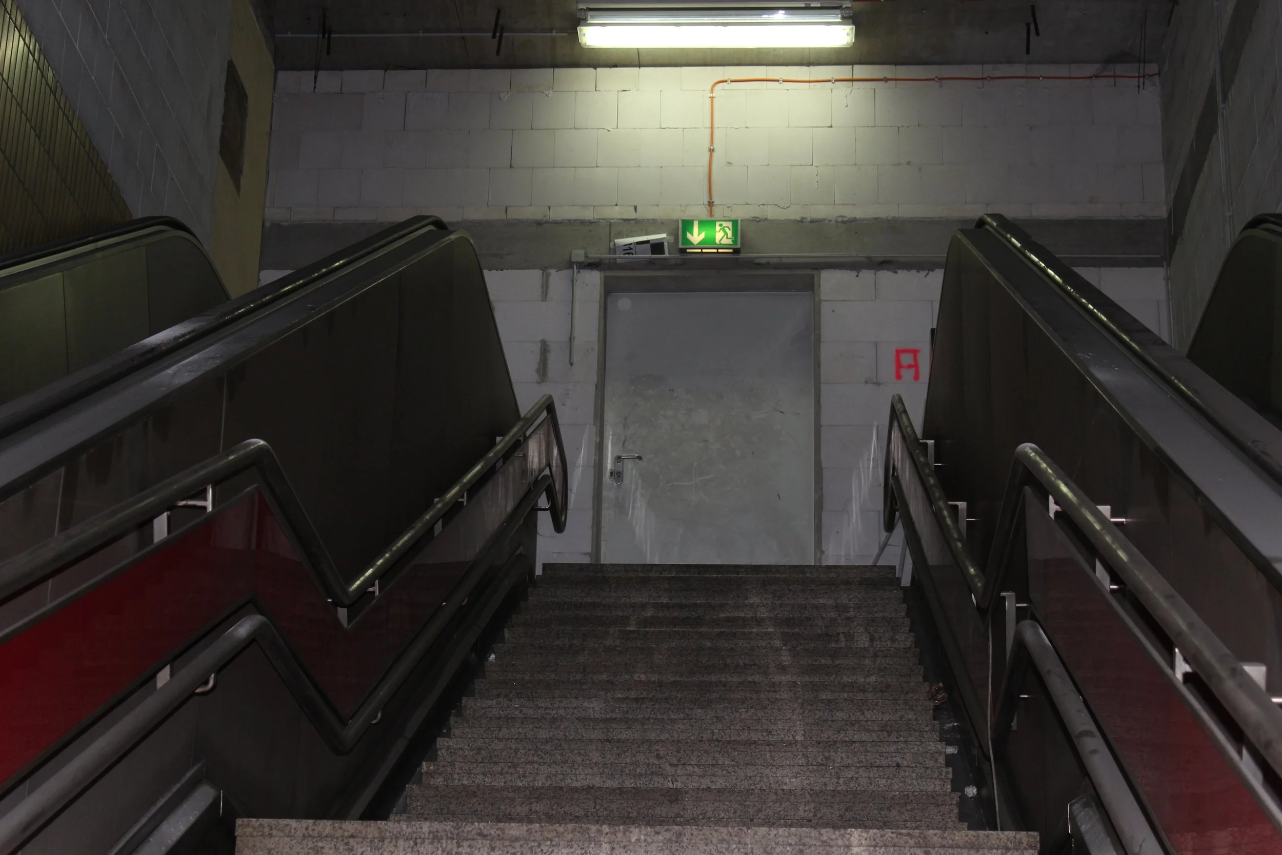 Es ist eine Treppe in einem unterirdischen Bereich mit einem Notausgangsschild über einer Tür, die nach oben führt. Die Umgebung ist unsauber und düster.
