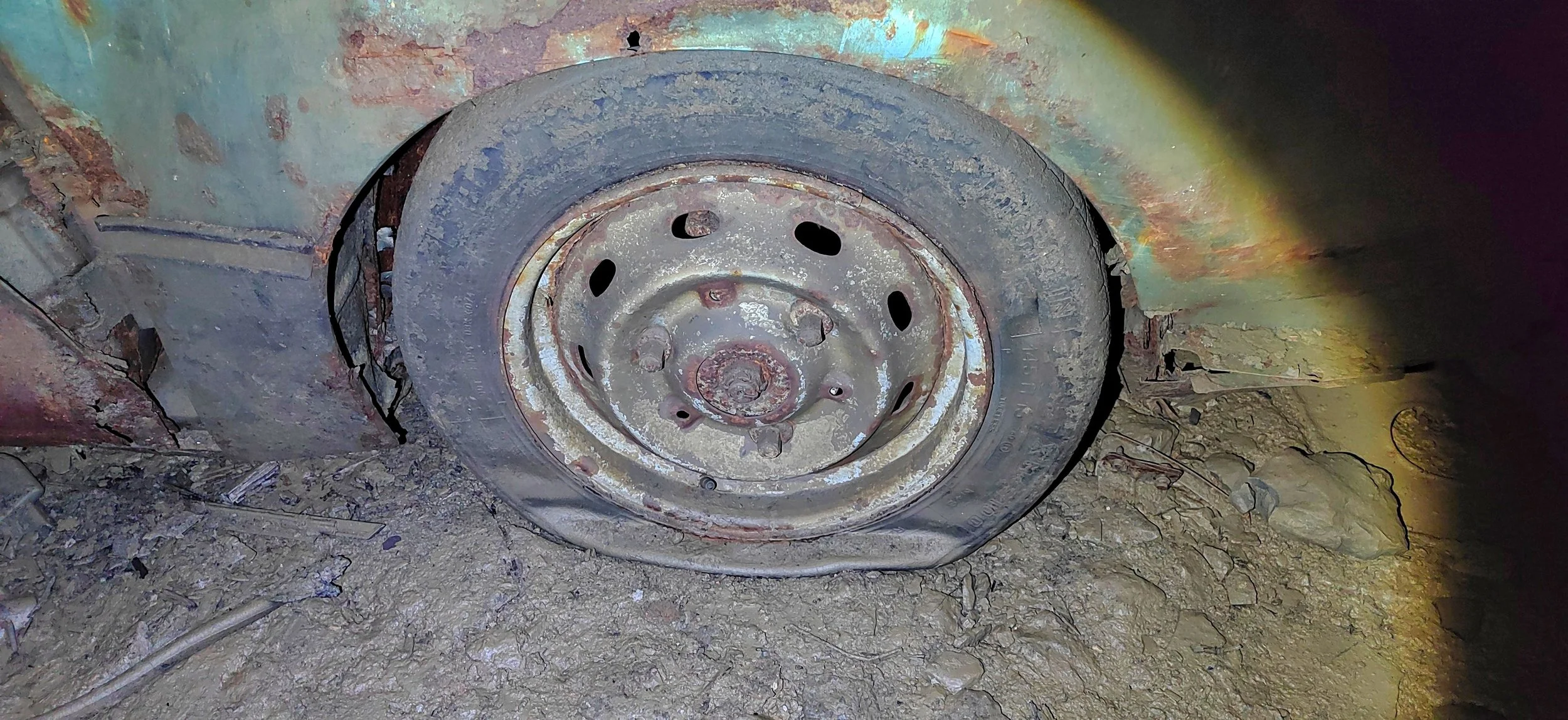 Verrosteter Reifen auf losem Boden, umgeben von Schmutz und kleinen Steinen, an einem alten und beschädigten Fahrzeug.