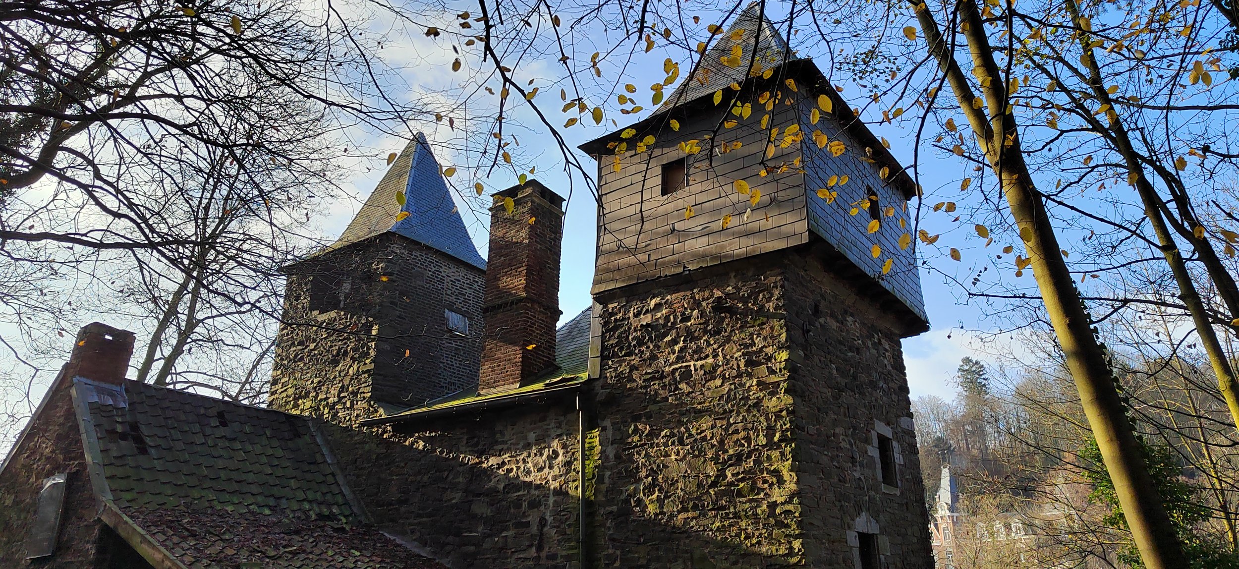 Alter Fachwerk- und Steinhäuser in einer Waldumgebung mit Bäumen, die im Herbst gelbe Blätter tragen. Das Bild zeigt einen blauen Himmel mit wenigen Wolken.
