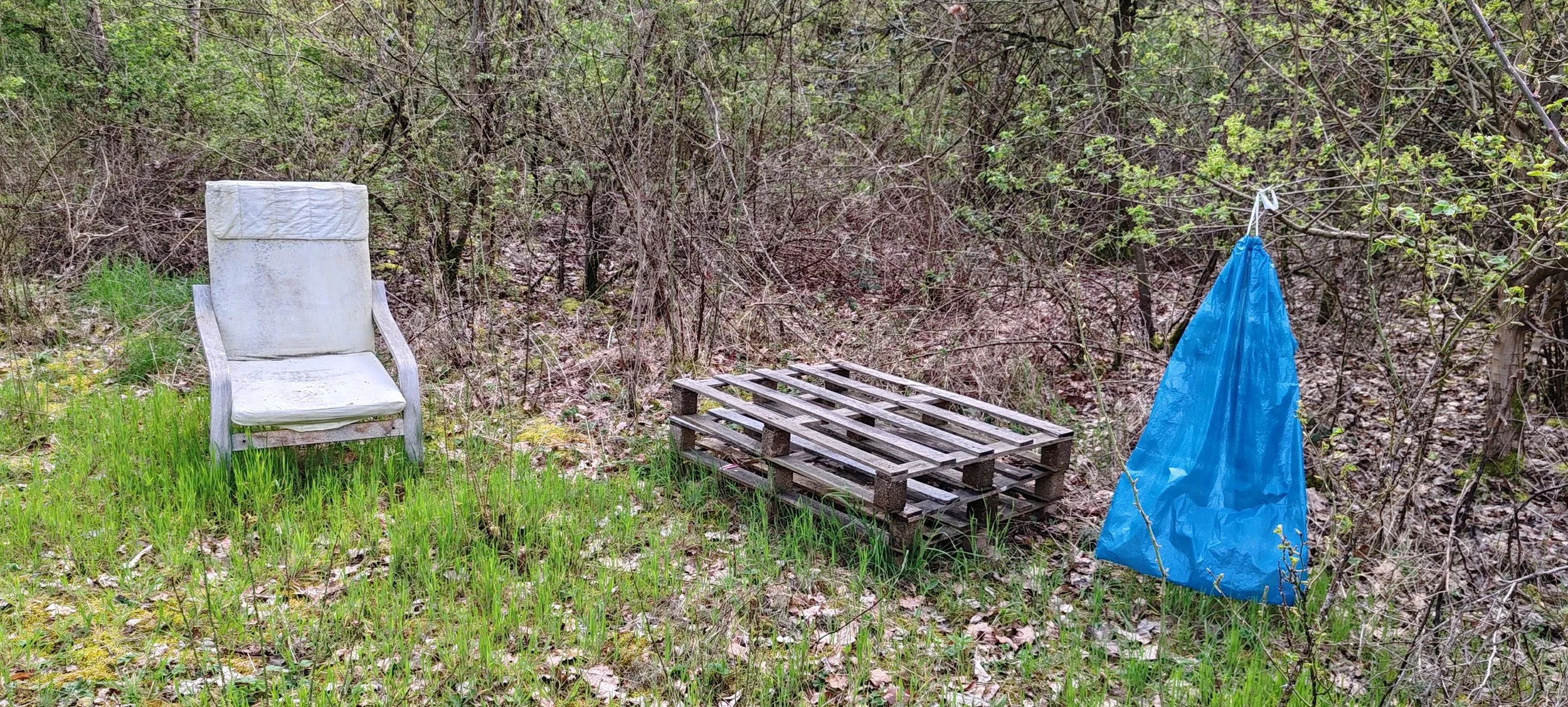 Ein alter, verrosteter Holzstuhl steht auf einer Wiese mit grünem Gras, im Hintergrund Gebüsch, daneben ein Palettenstuhl und ein blauer Plastiksack, der an einem Seil hängt.