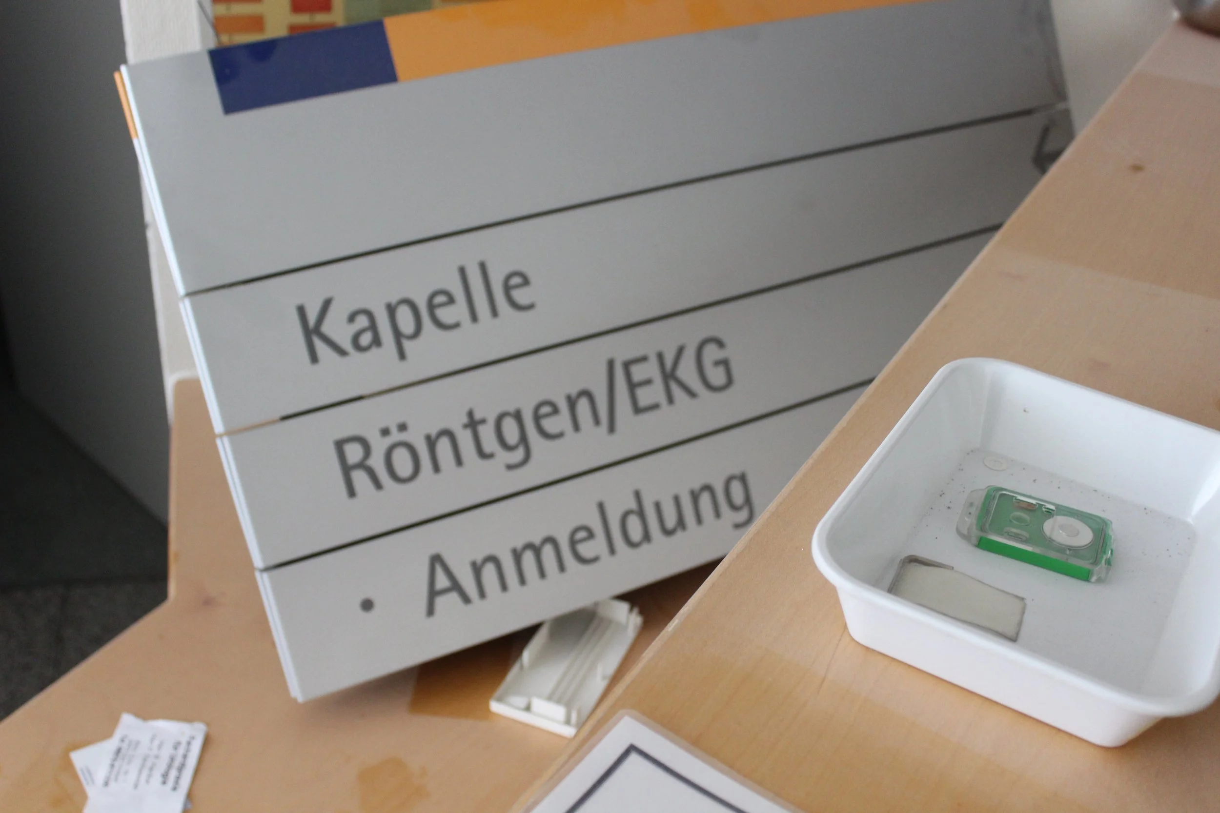 Ein Schild mit den Aufschriften 'Kapelle', 'Röntgen/EKG' und 'Anmeldung', sowie eine weiße Schale mit einem kleinen grünen Wasserwaagen darin auf einem Holztisch.