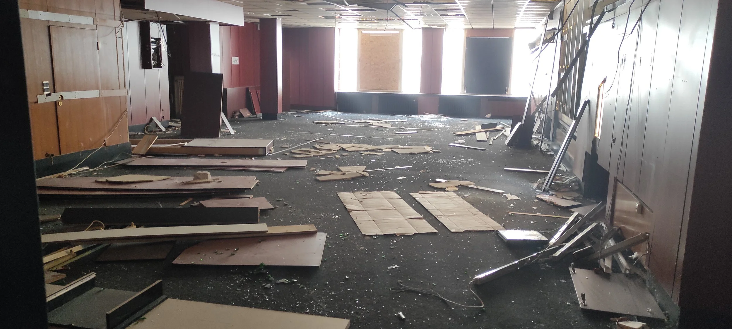 Innenraum eines zerstörten Gebäudes mit herumliegenden Trümmern, Papiertüchern, beschädigten Möbeln und zerbrochenen Fenstern.