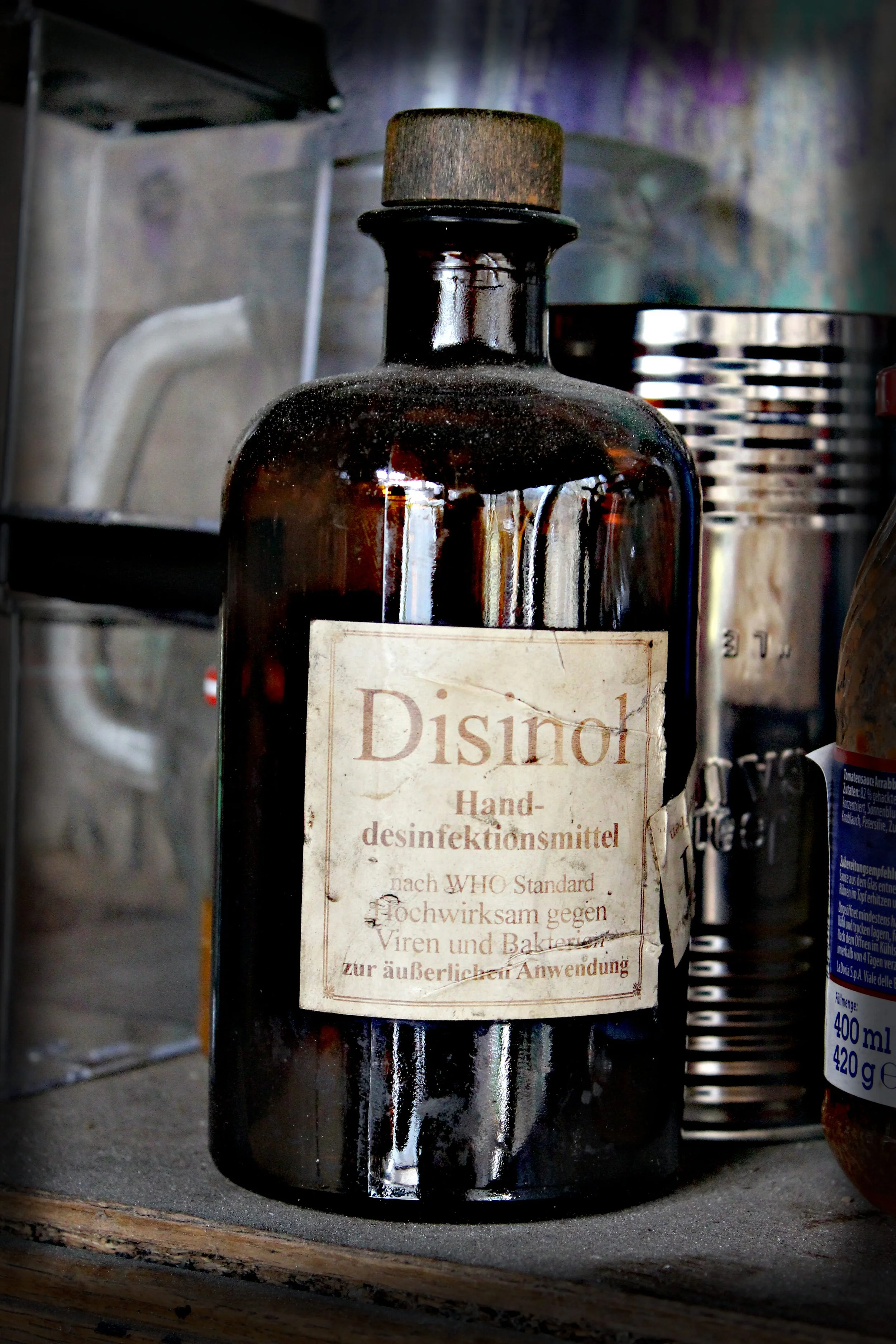 Ein brauner Glasflasche mit einem alten, vergilbten Etikett, das Handdesinfektionsmittel nach WHO-Standard beschreibt, auf einer Holzoberfläche neben weiteren Behältern.