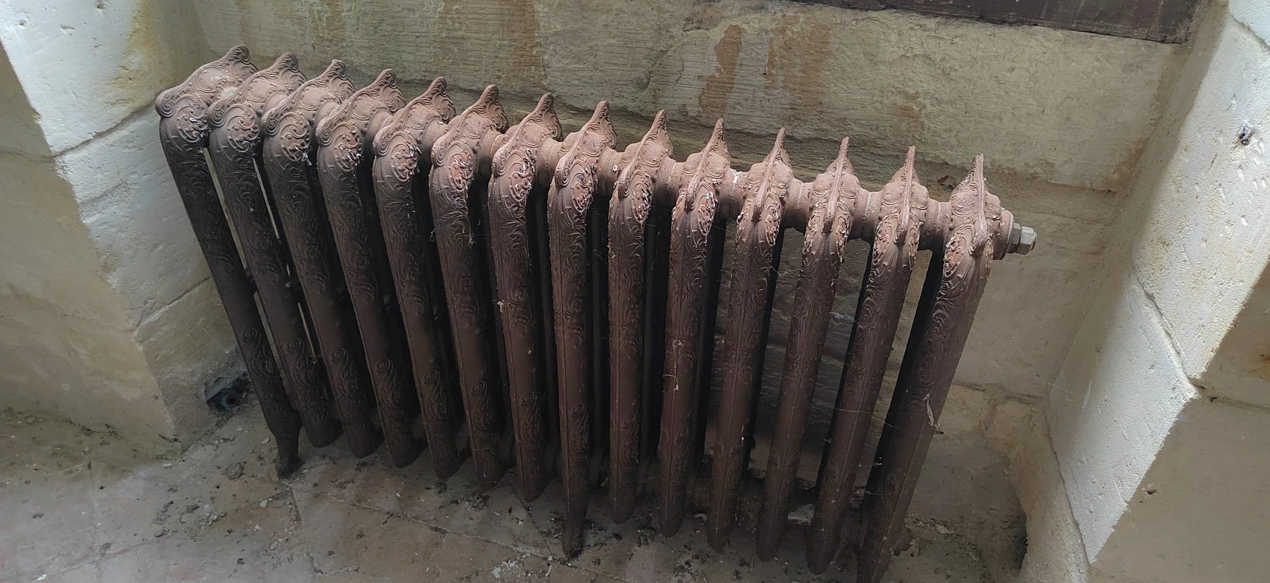 Alter, verrosteter Heizkörper mit kunstvollen Mustern in einem Raum mit Steinwänden.