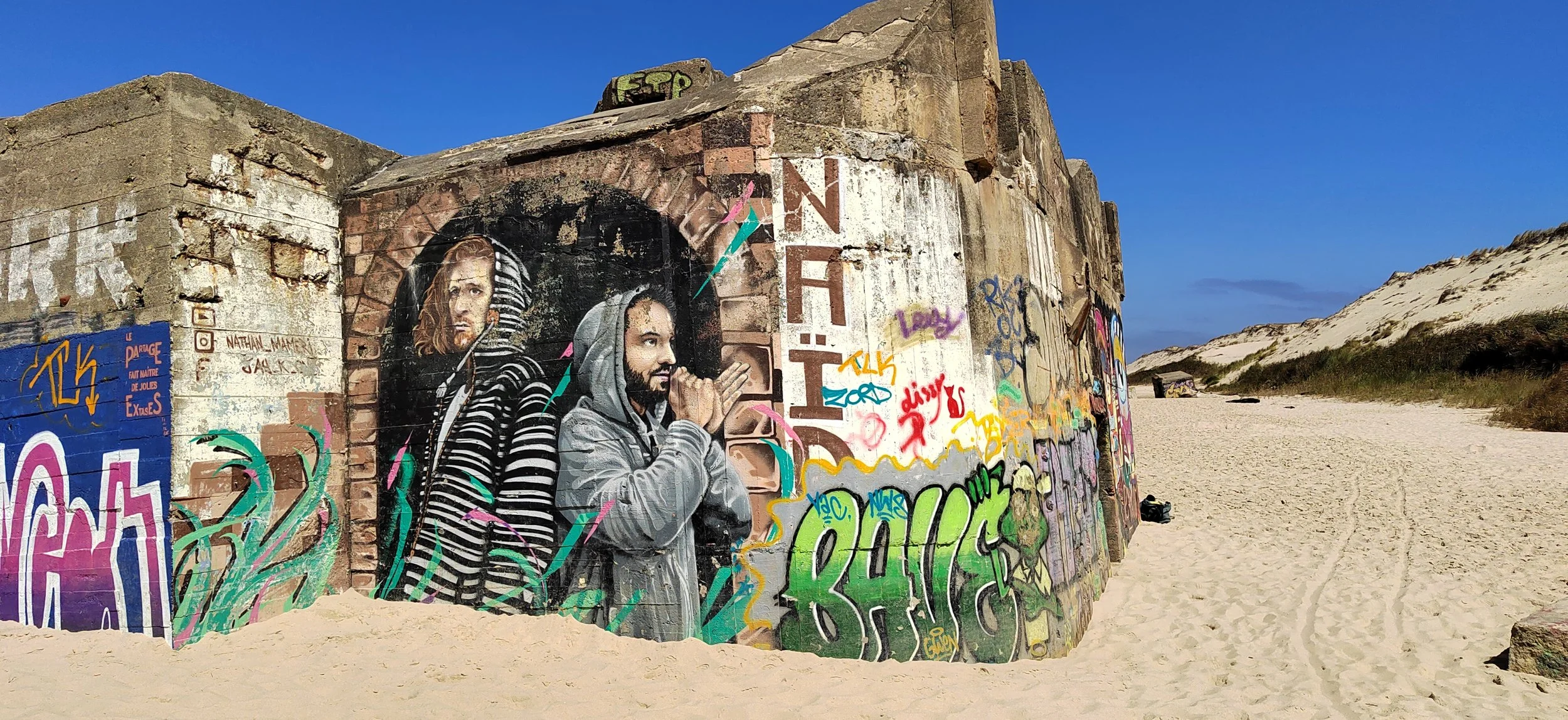 Graffiti-Kunst an einer Betonmauer in einer Dünenumgebung mit Sand, Gras und klarem blauen Himmel, zeigt zwei Männer, einer in Streifen-Kleidung, der andere mit Kapuze, in urbanem Stil, umgeben von bunten Tags und Schriftzügen.