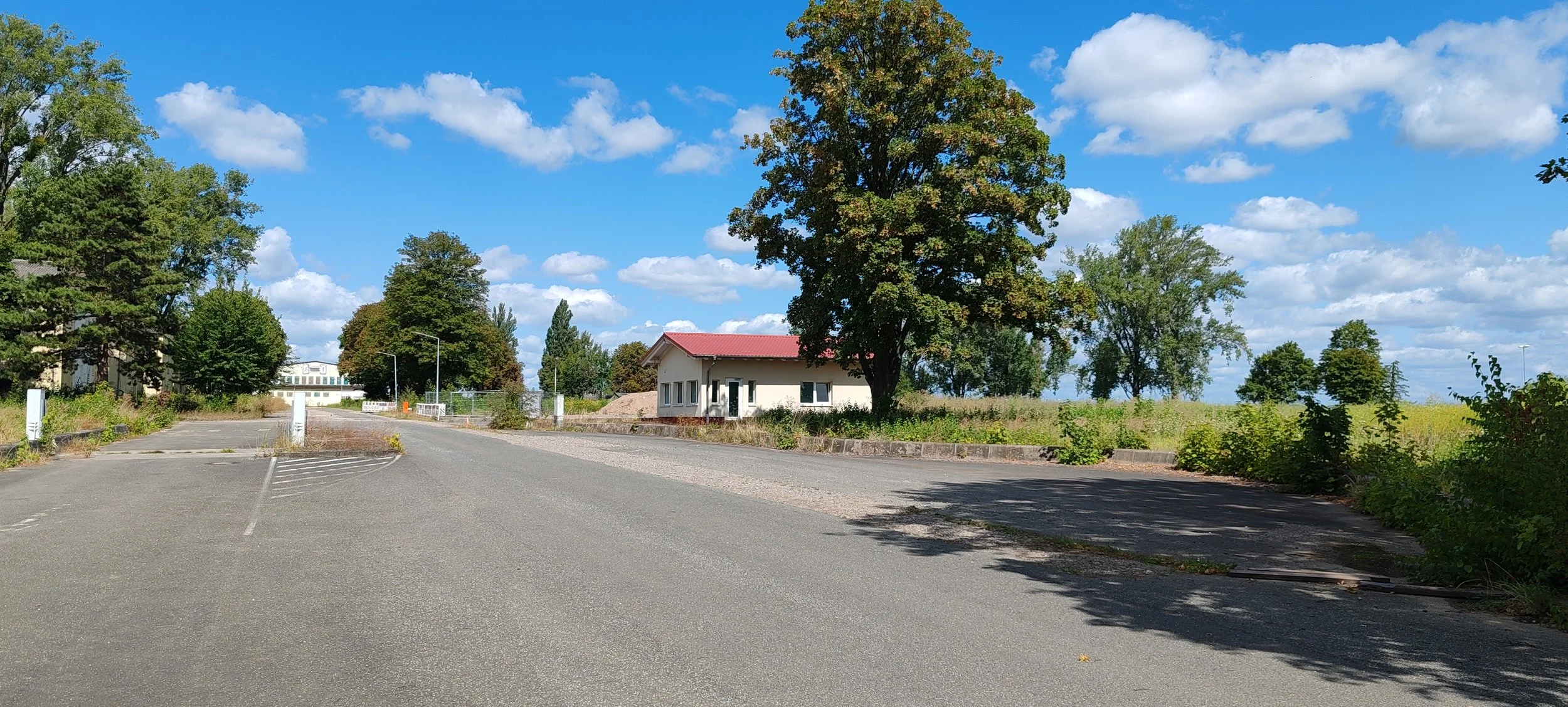 Ein leerer Parkplatz vor einem kleinen weißen Haus mit rotem Dach, umgeben von Bäumen und einer grünen Wiese unter einem blauen Himmel mit Wolken.