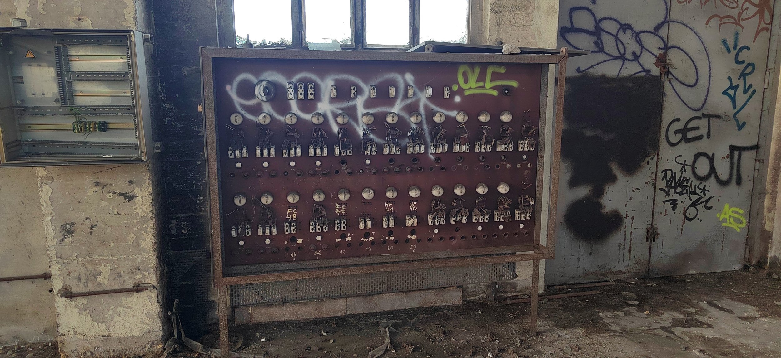 Ein verrosteter, alter elektrischer Verteilerkasten mit offenem Deckel und einem grauen, vandalisierten Schalteinstrument in einem verlassenen, beschmutzten Raum, mit Graffiti an den Wänden.