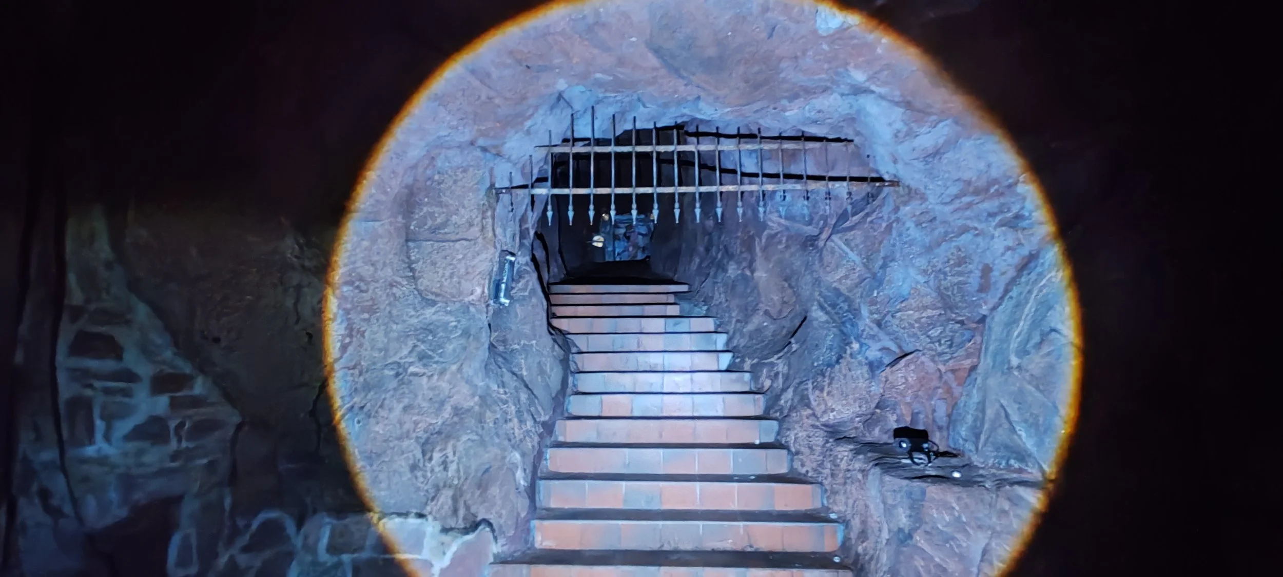 Blick in einen Höhleneingang, durch den eine Treppe nach oben führt. Die Höhle ist aus Fels mit unregelmäßiger Oberfläche. Es befinden sich zwei kleine Gegenstände, möglicherweise Taschenlampen, am Boden rechts.