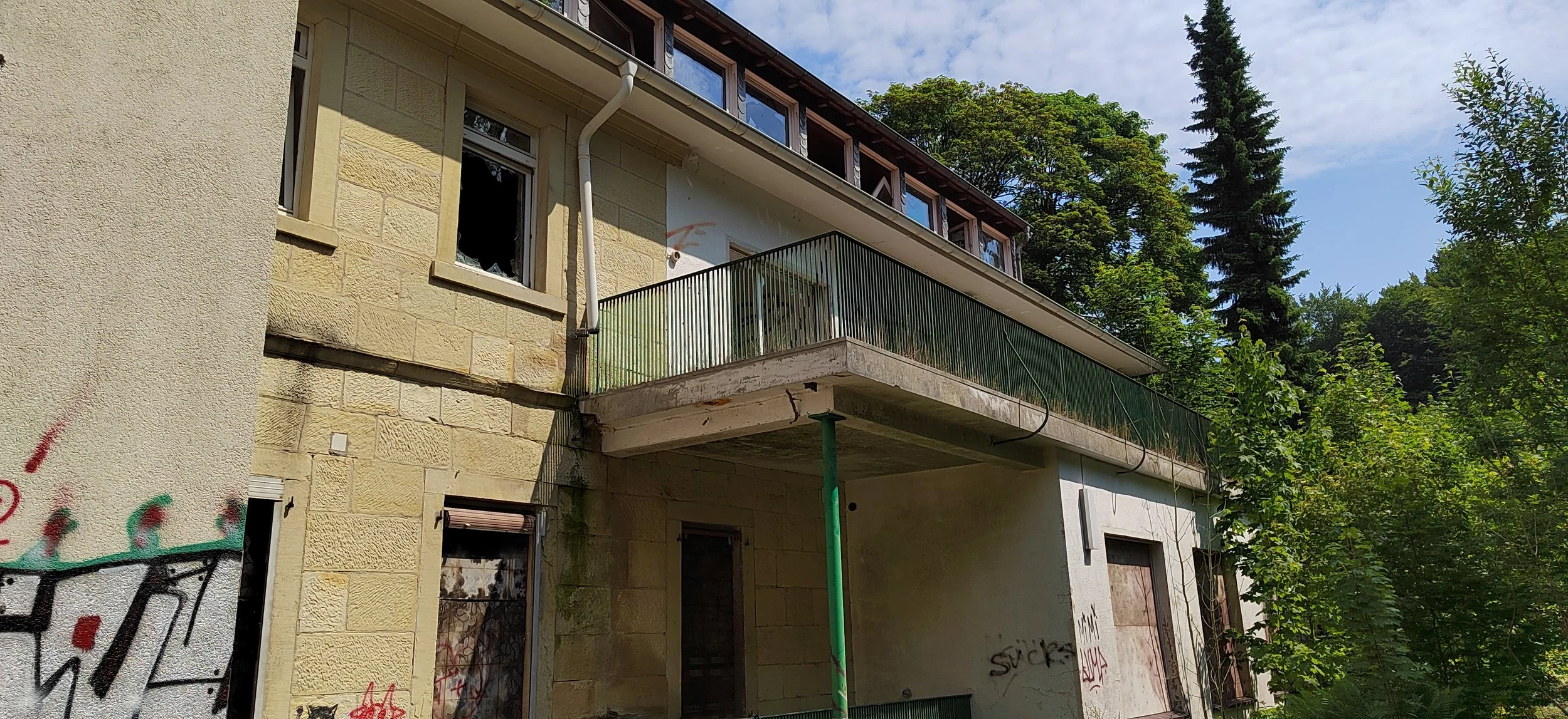 Verfallenes Haus mit Graffiti, beschädigten Fenstern, einer Betonbalustrade auf einem kleinen Balkon und umgeben von Bäumen und Natur.