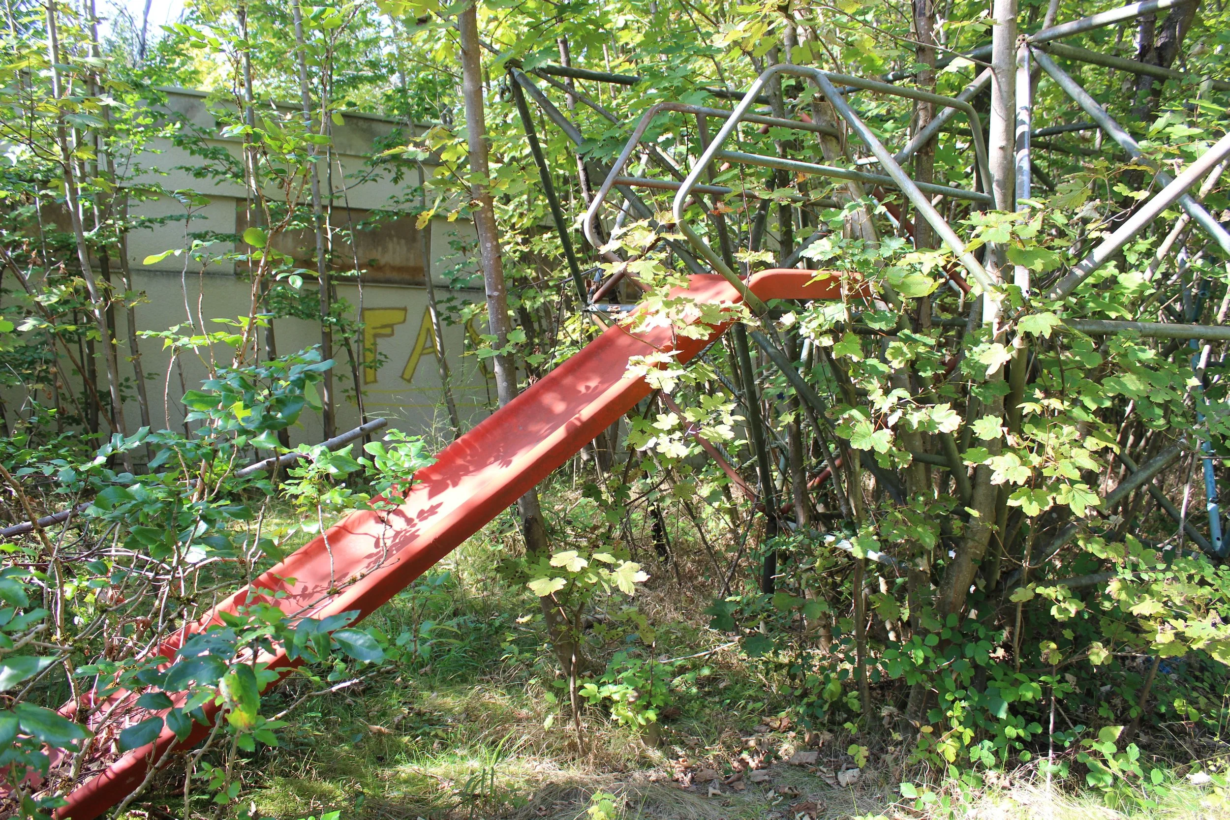 Ein verrosteter roter Spielplatzrutschentunnel, überwuchert von grünen Büschen und Bäumen, im Freien.