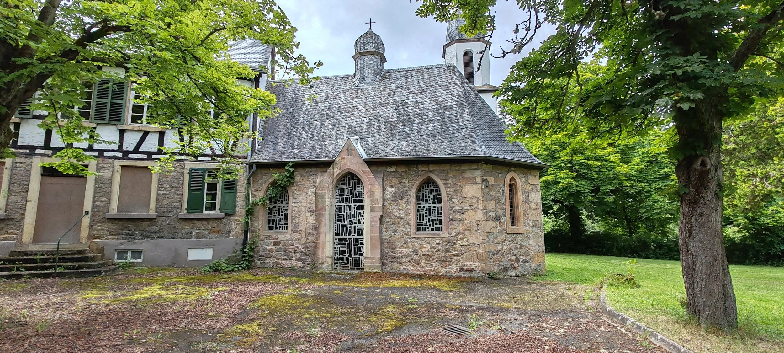 Kleine romanische Kirche mit Steinmauer, schiefergedecktem Dach und buntem Fenster, am Rande eines Baumes. Umgebung mit grünen Bäumen und Gras.