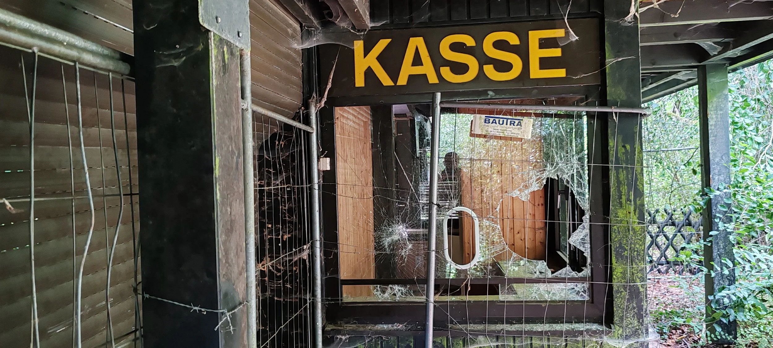 Zerstörtes Kassenhäuschen mit einem zerbrochenen Glastür und beschädigtem Metallzaun, über dem steht das Wort 'KASSE' in großen gelben Buchstaben, umgeben von Spinnweben und Vegetation.