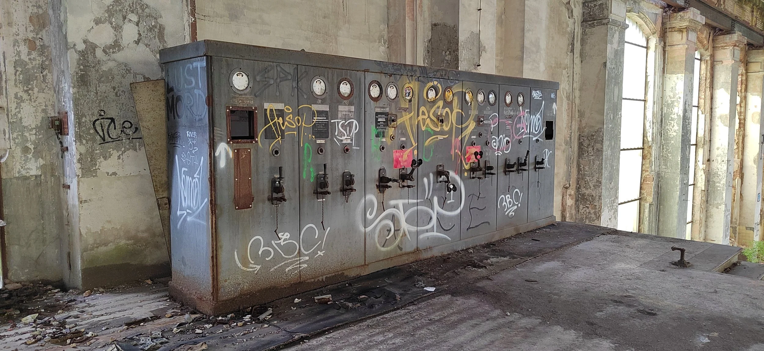 Ein verrosteter und mit Graffiti besprühter alter Stromverteilerkasten in einem verfallenen, ungepflegten Gebäude mit großen Fensteröffnungen.