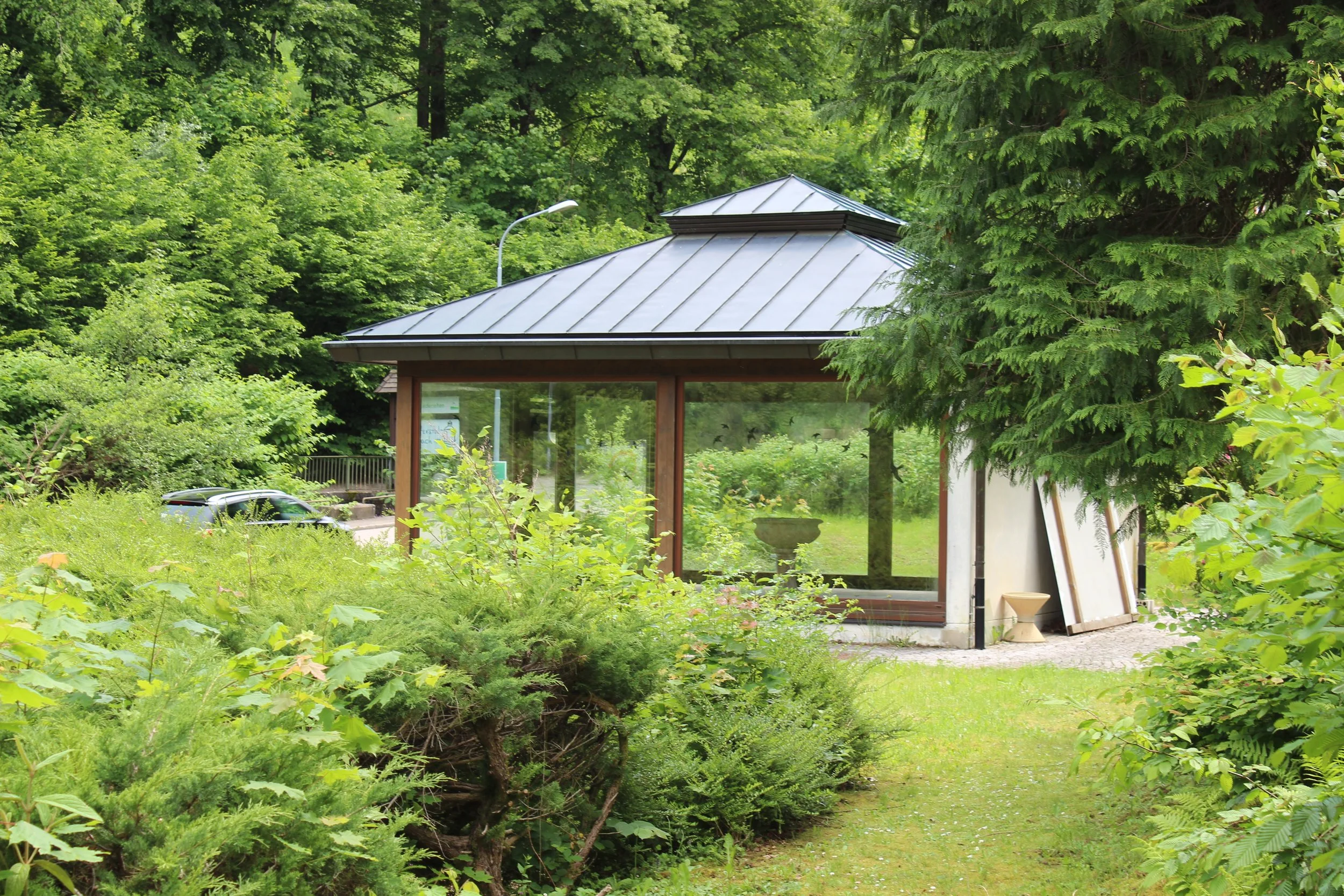 Gemütlicher Glashaus inmitten grüner Natur, umgeben von Büschen und Bäumen, mit Blick auf einen Garten und einen Parkplatz im Hintergrund.