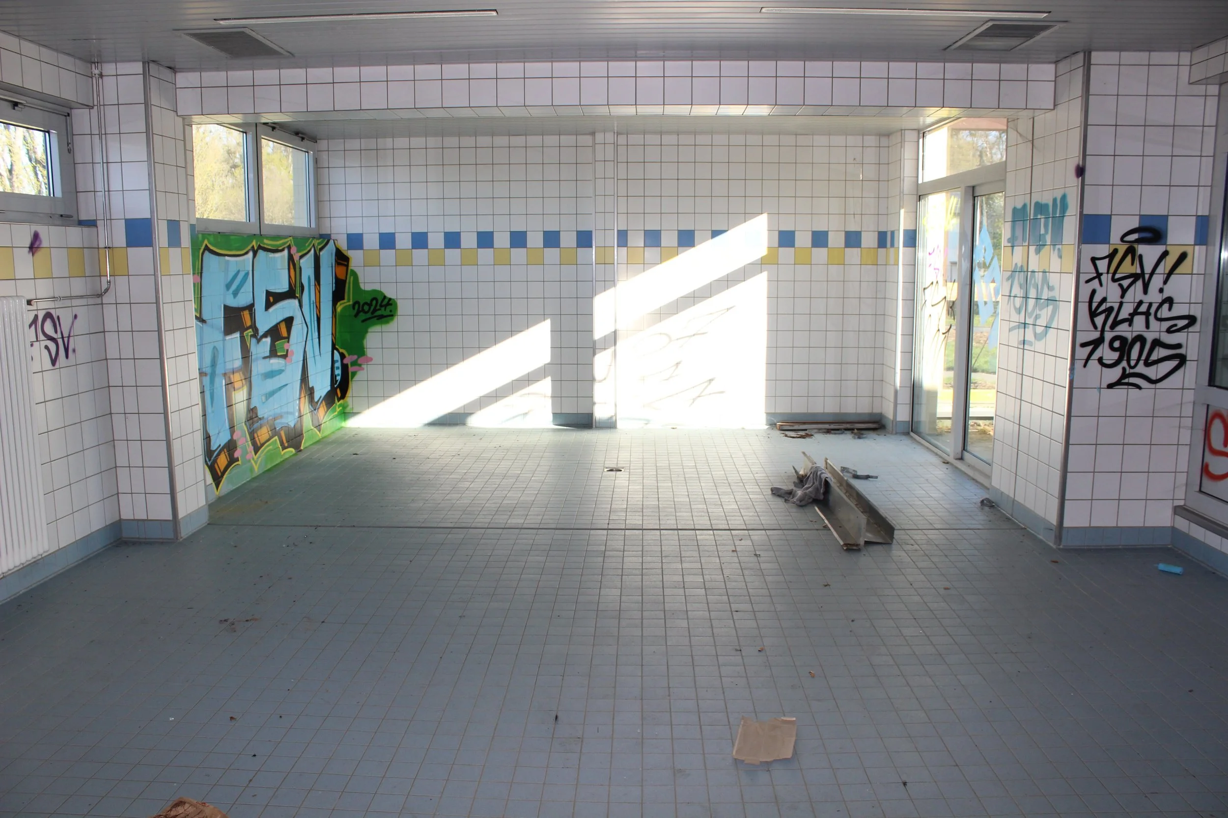 Leerer Raum mit weißen Wandfliesen, Grautreppe, Grautüren und Graufliesenboden, bemalt mit Graffiti, Sonnenlicht und Schatten an den Wänden.