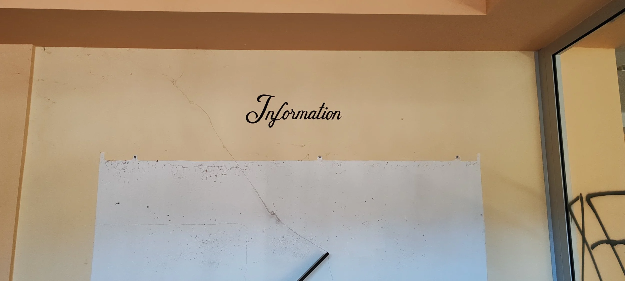 Ein Wandposter mit der Aufschrift 'Information' über einem weißen Brett in einem Raum mit brauner Wand und Fenstern. Das Poster ist leicht beschädigt.
