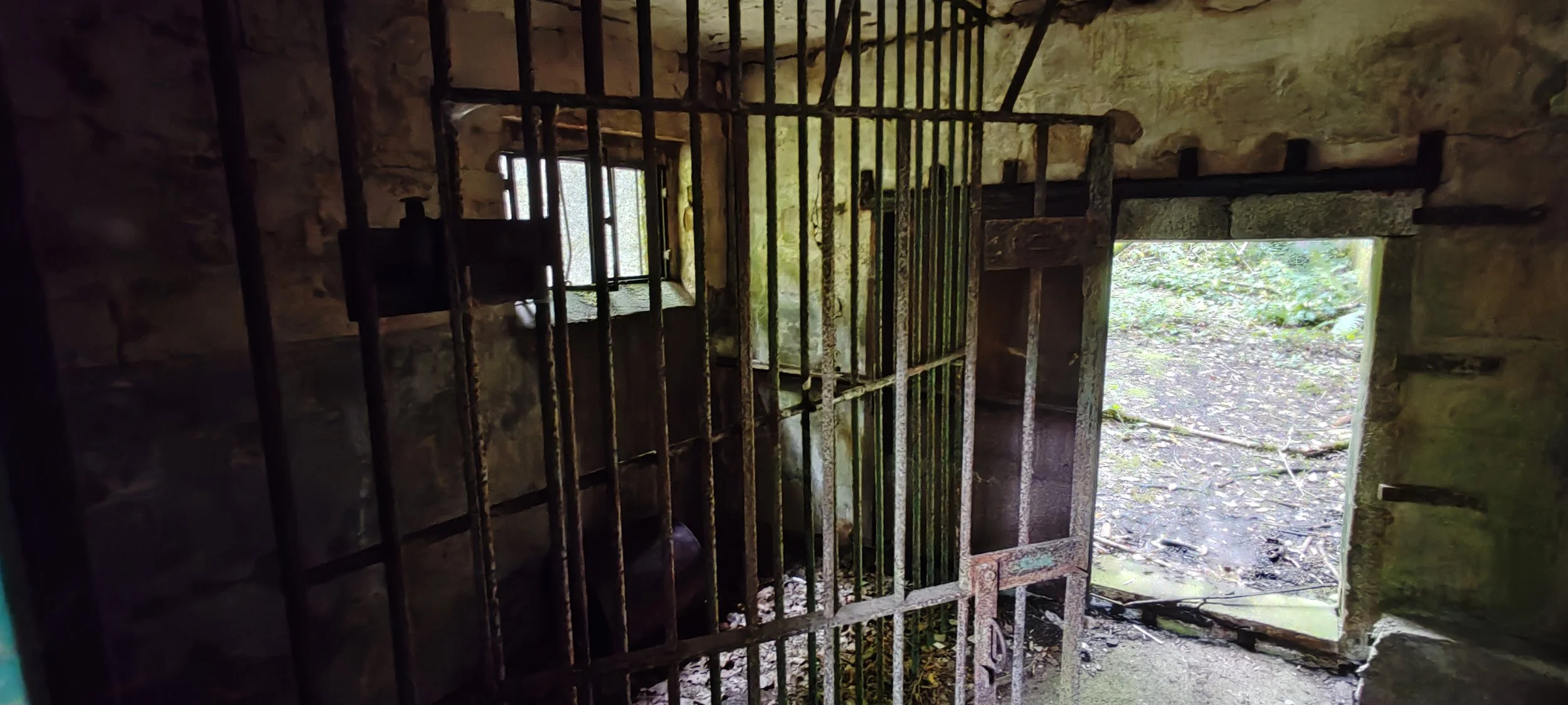 Verrostete Gittertür in einer alten Stein-Hütte, offene Tür führt nach draußen in den Wald, Sonnenlicht scheint durch das Fenster und den Türspalt.