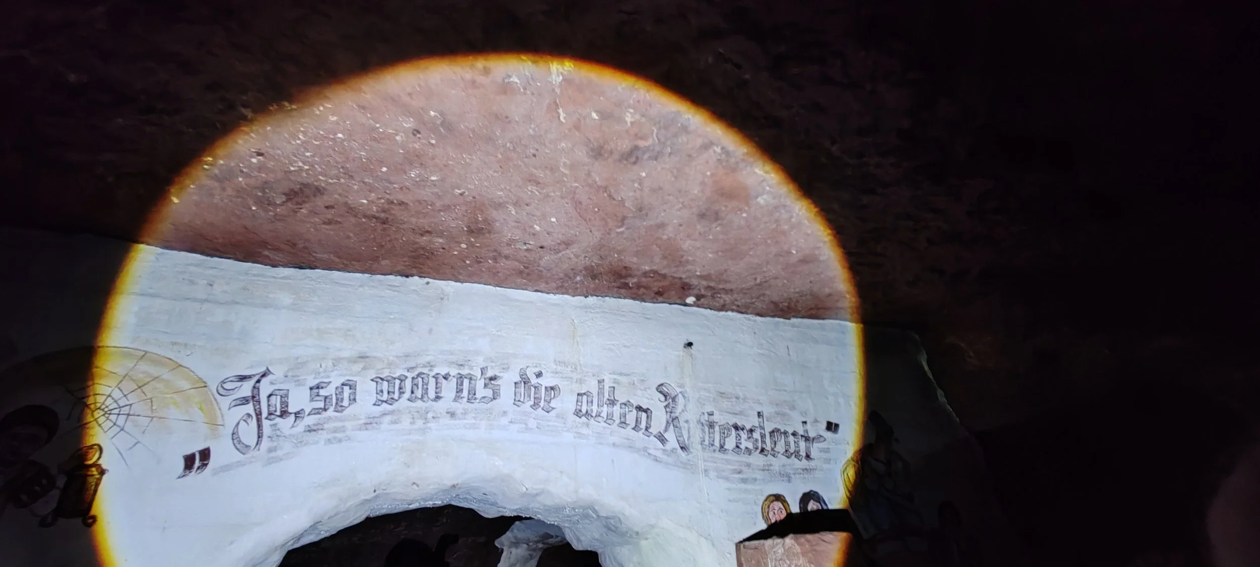 Innenansicht eines Höhleneingangs mit Wandgemälde, das in deutscher Sprache eine Geschichte erzählt, mit einem Lichtstrahl, der den Raum beleuchtet.