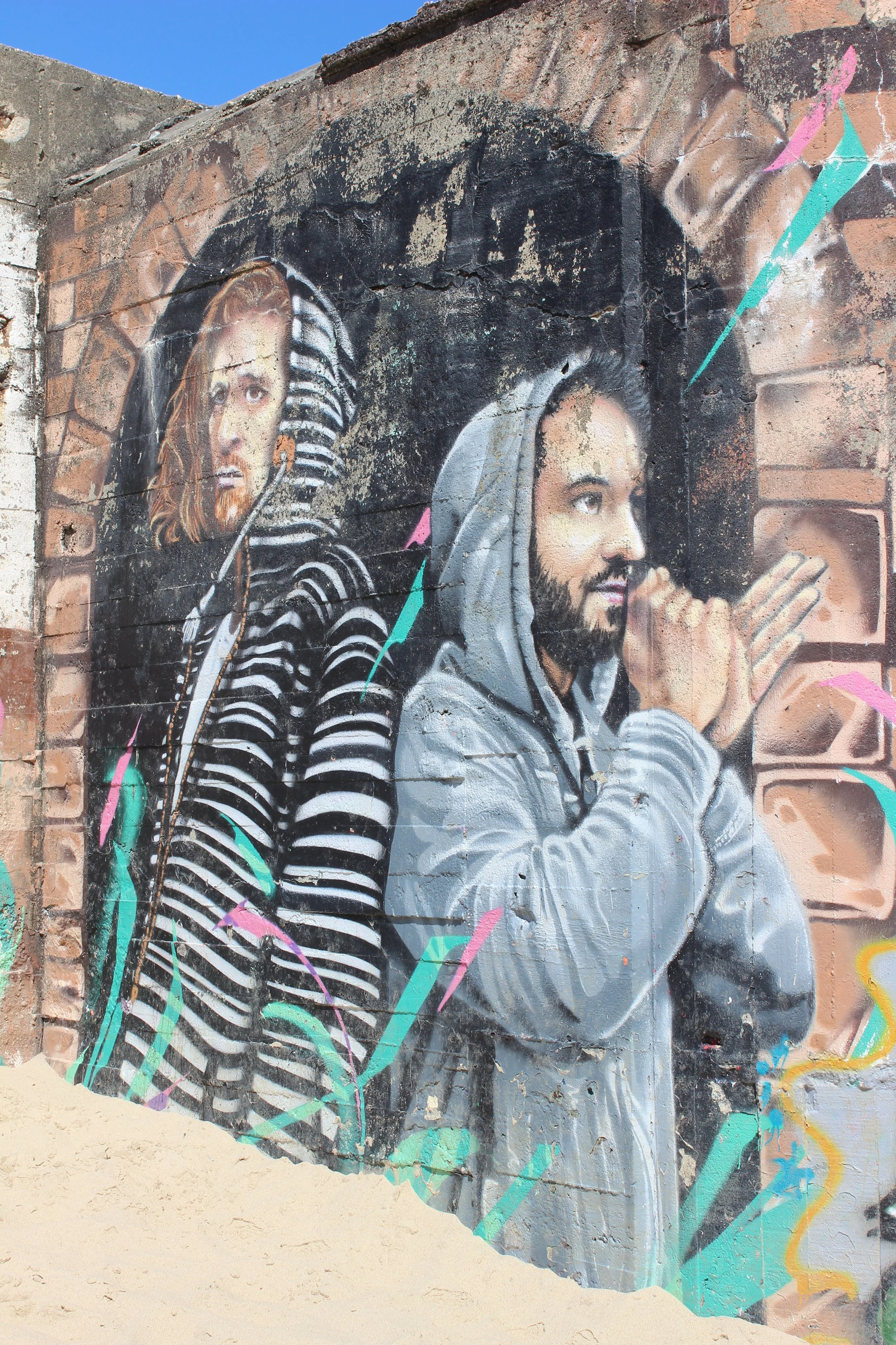 Straßenkunst mit zwei männlichen Figuren, einer trägt ein Zebra-Strickpullover und einen Kopfhörer, der andere trägt einen grauen Hoodie und hat die Hände vor dem Gesicht. Farbenfrohe Akzente und abstrakte Linien umgeben die Wand.