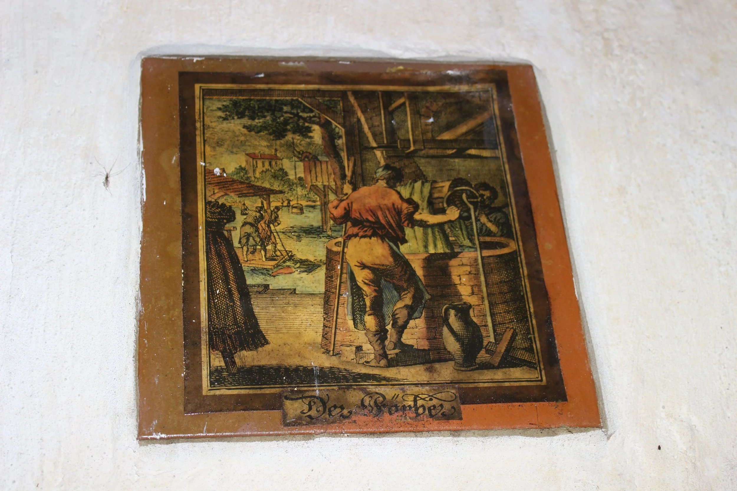 Alte Wandfliese mit einer Illustration eines Handwerkers bei der Arbeit, im Hintergrund ein Fluss und Menschen, darunter ein Schild mit der Aufschrift "Der Weber".