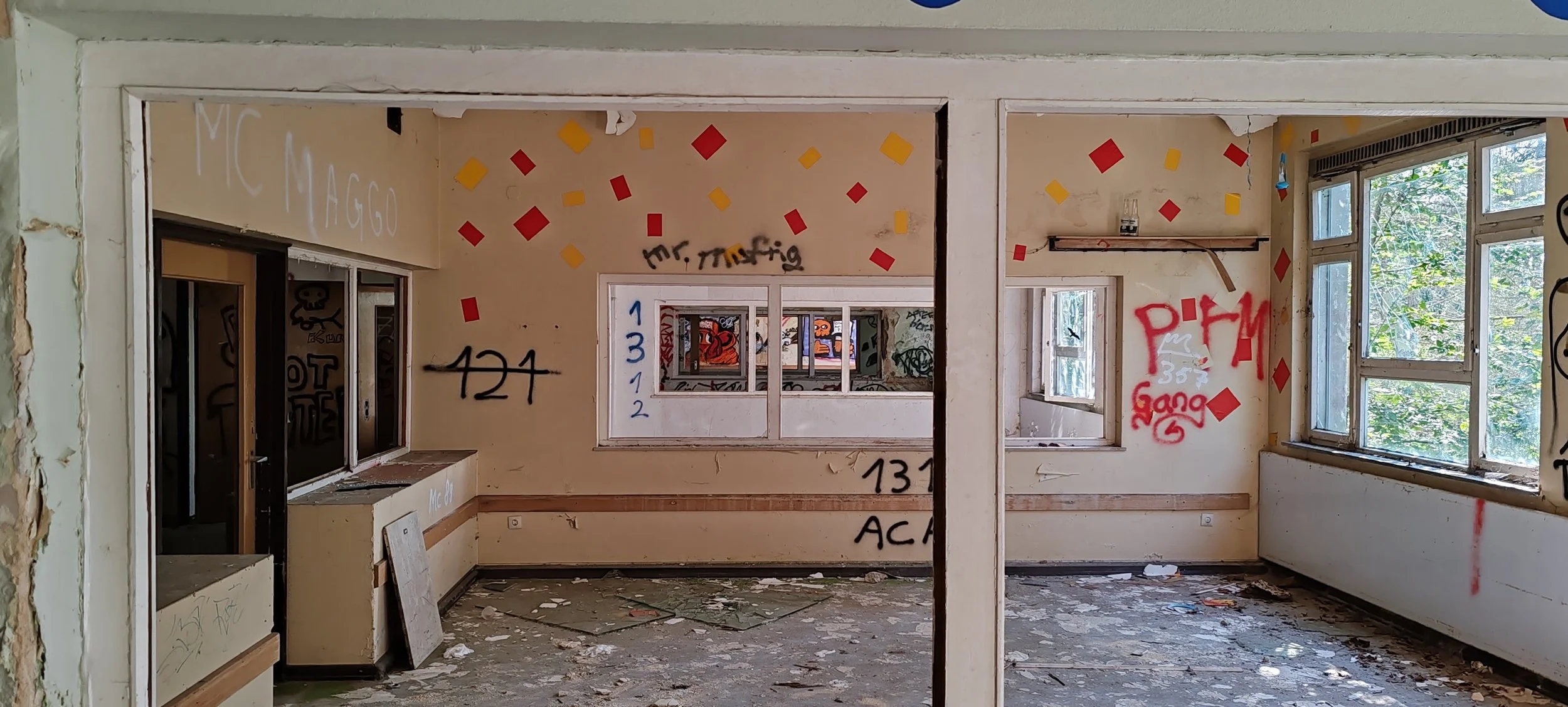 Innenraum eines verlassenen, grauen Gebäudes mit zerbrochenen Fenstern, grauem Boden und voller Graffiti auf den Wänden.