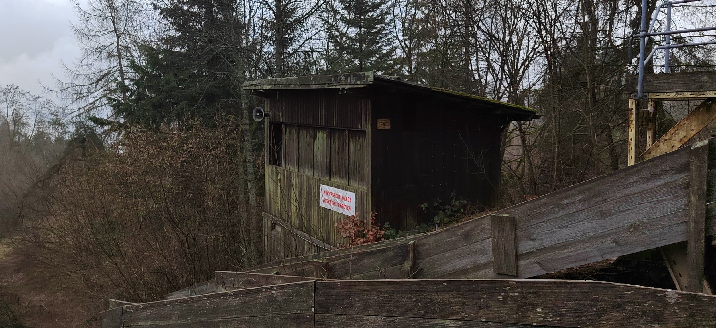 Ein dunkler Holzschuppen in einem bewaldeten Gebiet mit einem roten und weißen Schild an der Seite, umgeben von Bäumen und gebrochener Holzstufen.