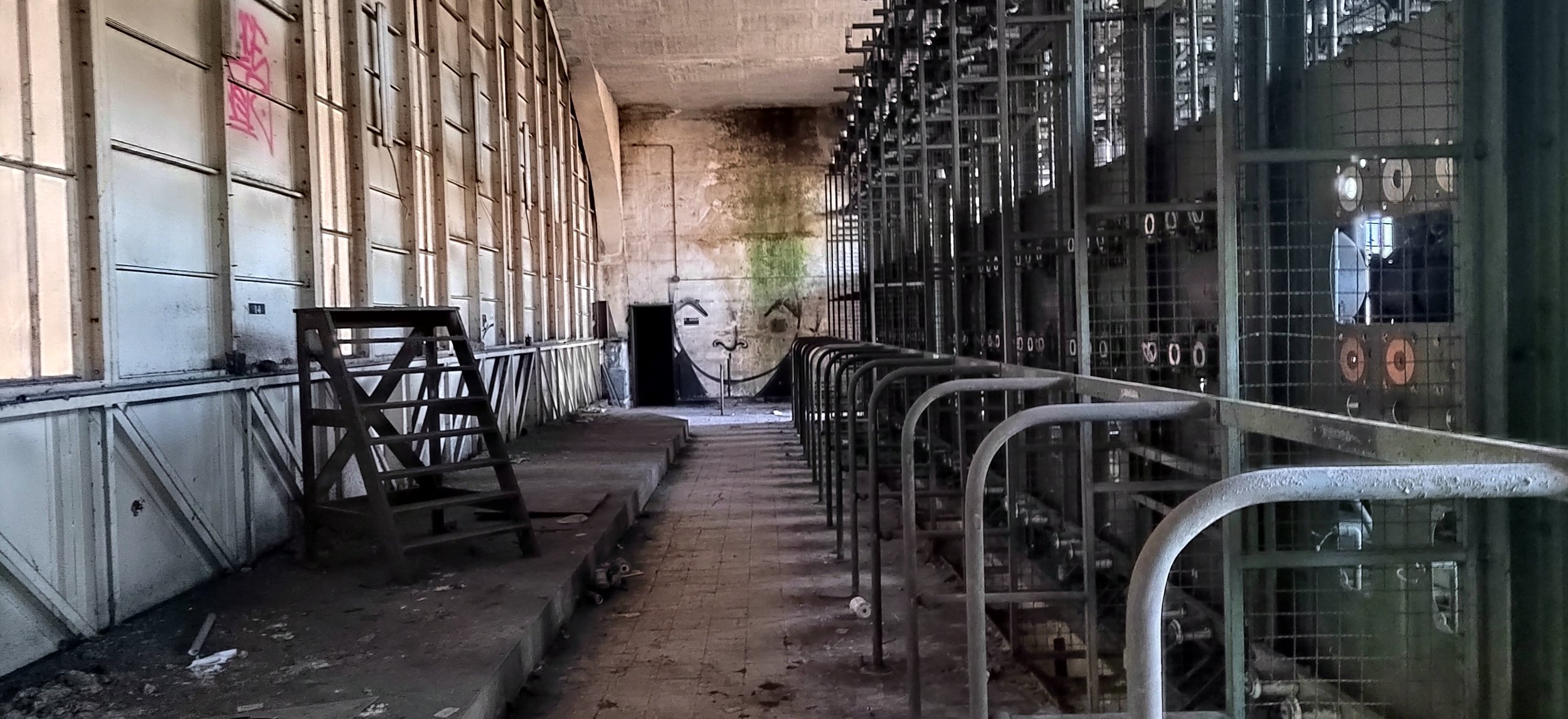 Verlassene industrielle Raum mit zerbrochenen Fenstern, verrosteten Regalen und Müll am Boden, Blick durch die Lagerhalle nach hinten, überwiegend grau und grün.