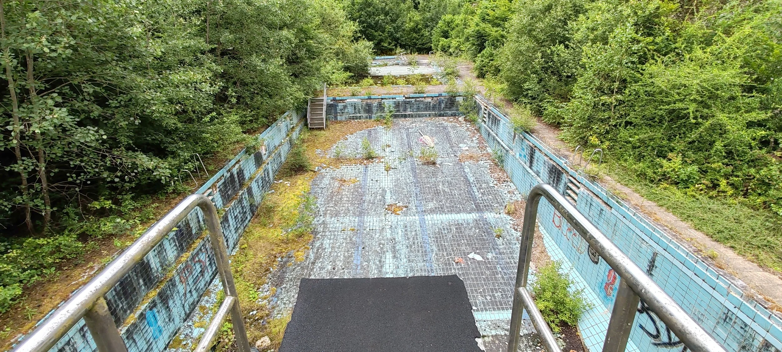 Verlassenes, verrostetes Schwimmbad im Freien, überwuchert von Pflanzen, mit Rasen und Bäumen im Hintergrund.