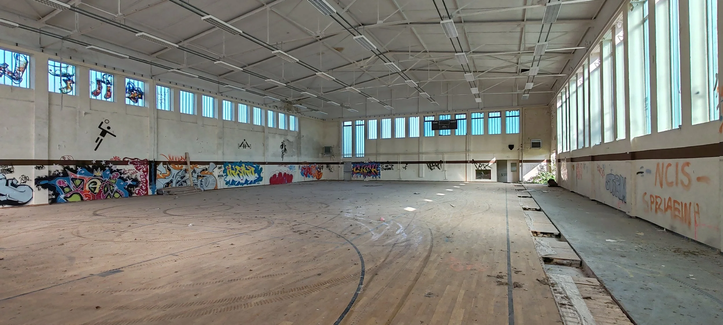 Leeres, verlassenes Hallenbad mit Graffiti an den Wänden, große Fenster an der Seite, ein Basketballkorb an der Wand, und verrosteter Bodenbelag.