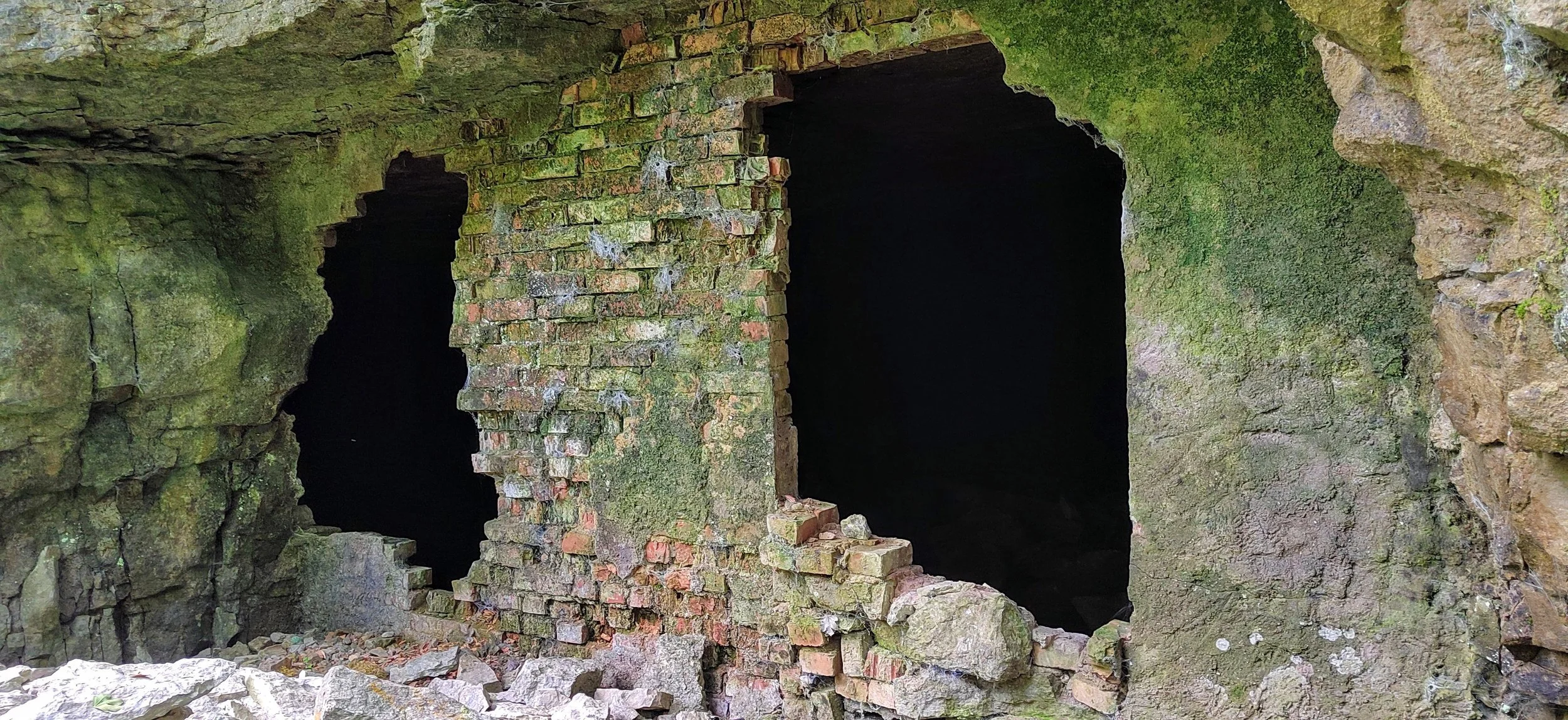 Alte, moosbedeckte Stein- und Ziegelmauer mit zwei rechteckigen Öffnungen, im Inneren dunkel und unzugänglich, umgeben von Trümmern und Natur.
